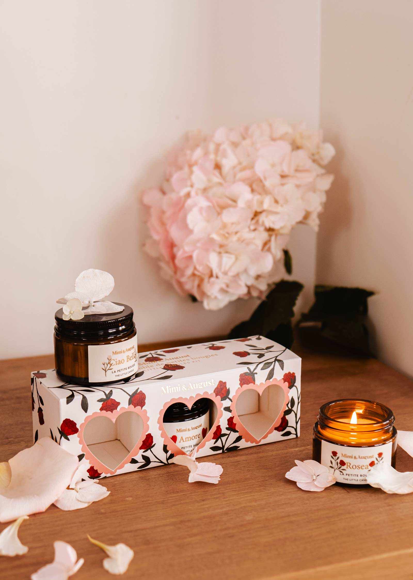 Two elegant candles in glass jars, one lit, adorn a wooden surface beside the Mimi & August La Romance Box Set. Exclusive fragrances fill the air as pink hydrangeas and scattered petals add floral charm to the scene.
