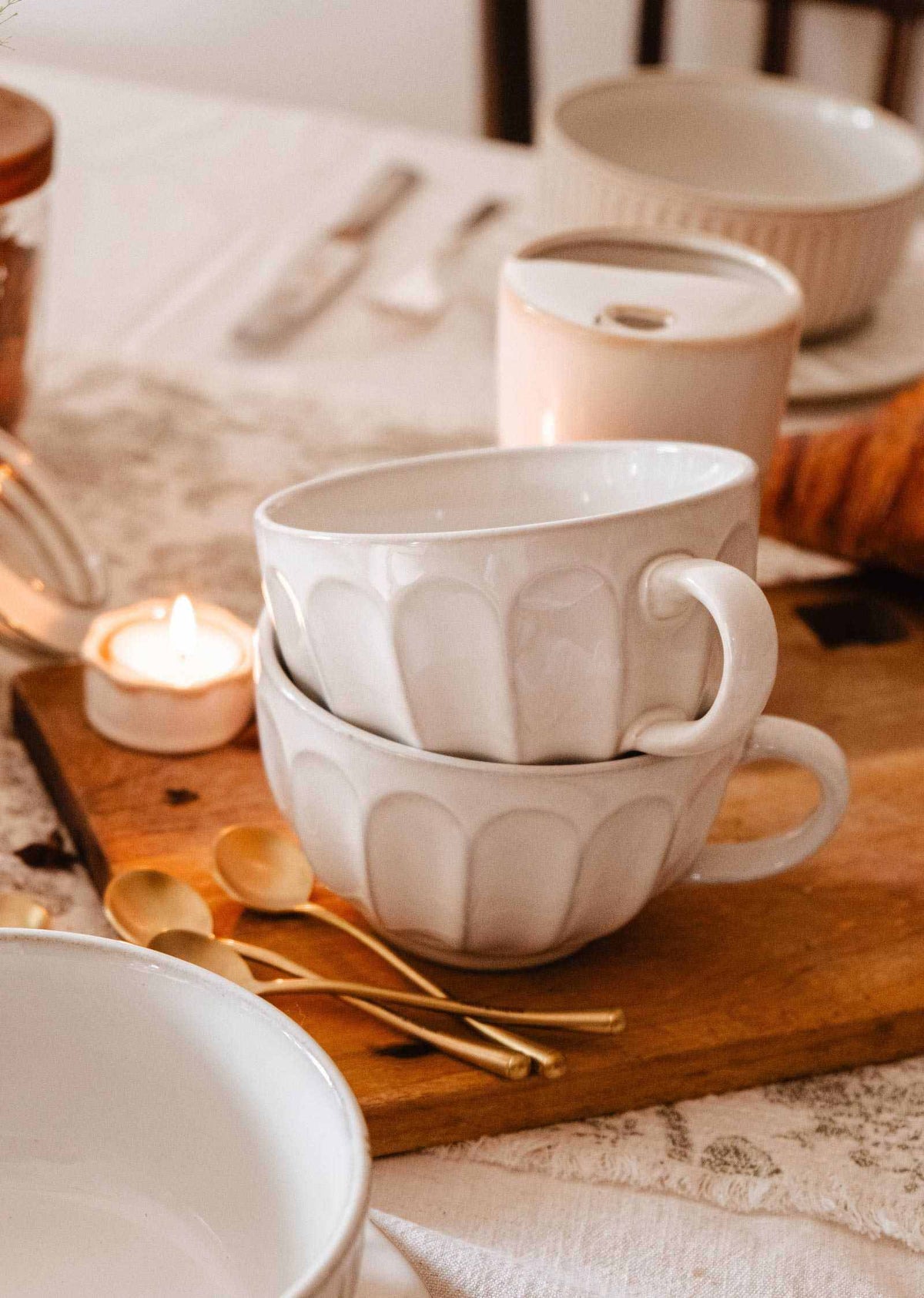 Two stacked Large Ceramic Latte Mug by Mimi & August sit on a wooden board beside gold spoons, a lit tealight candle, and other tableware on a white tablecloth.