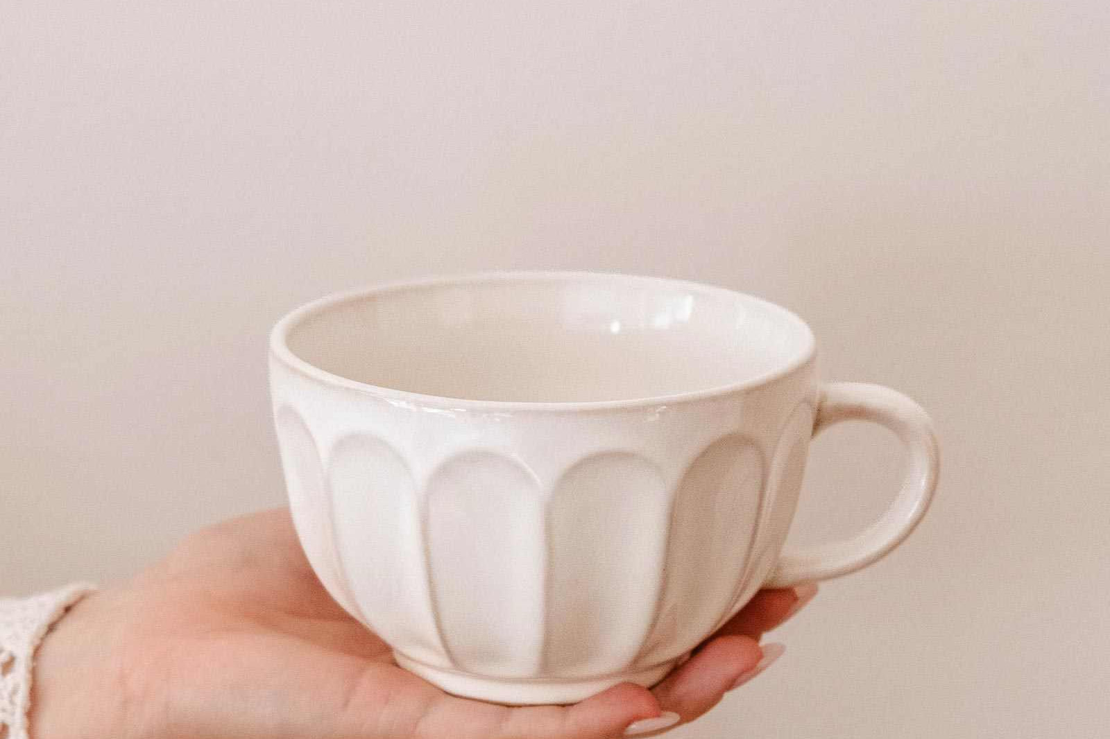 A hand holds a Ceramic Latte Mug by Mimi & August, displaying its white ceramic body with a textured, scalloped pattern against a plain light background.