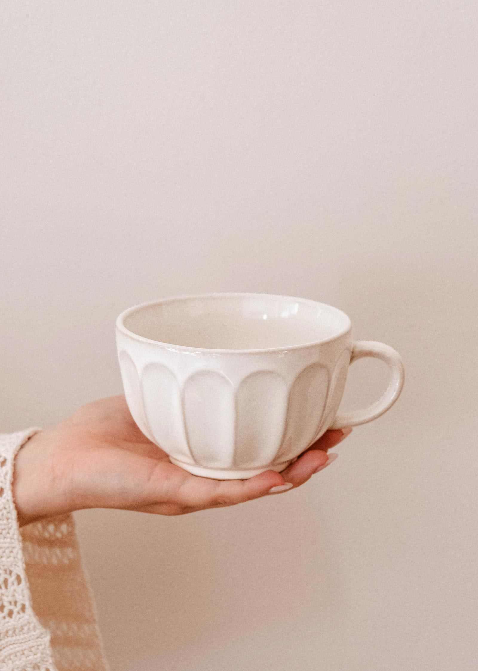 A hand holds a Ceramic Latte Mug by Mimi & August, displaying its white ceramic body with a textured, scalloped pattern against a plain light background.