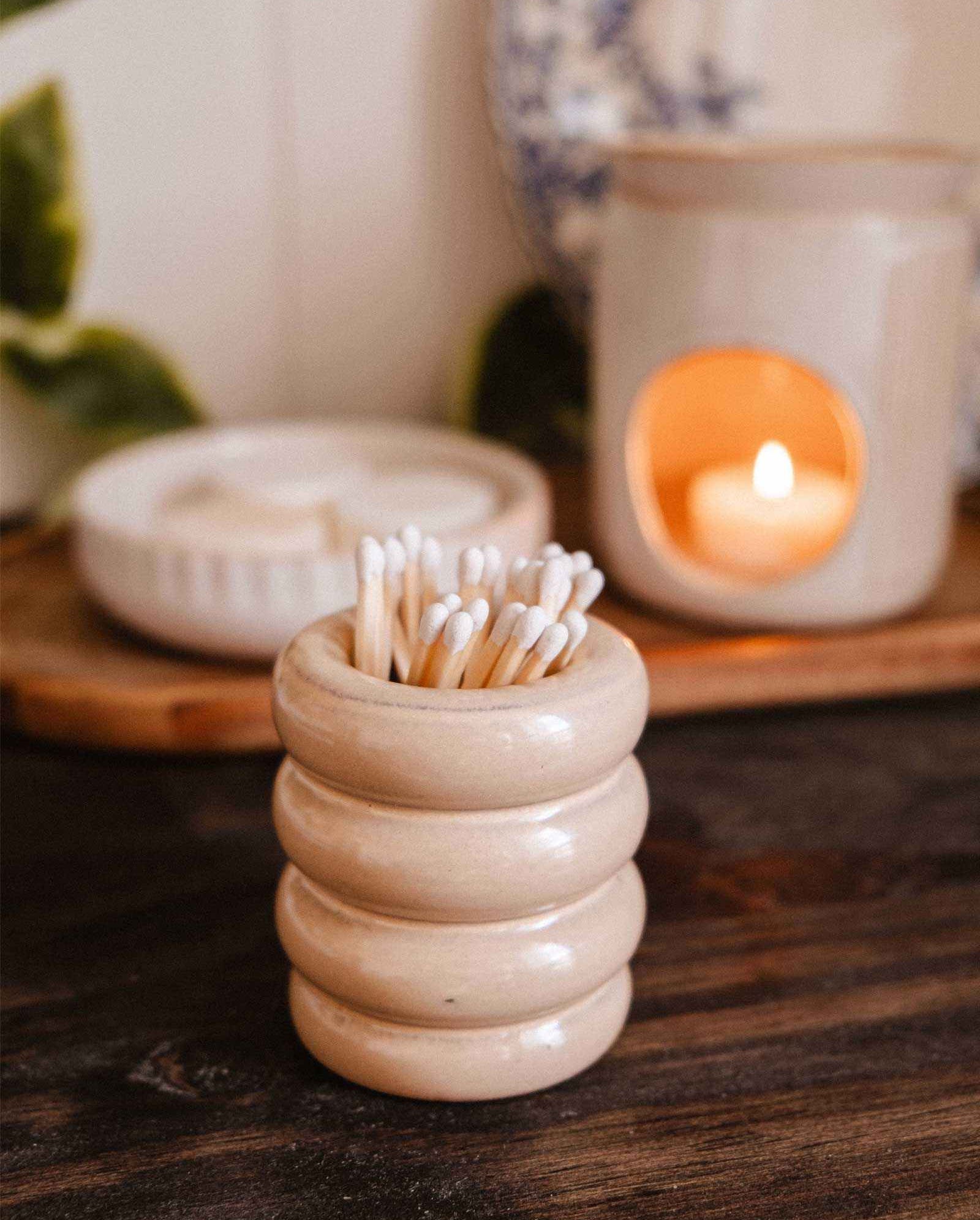 The Mimi & August Ceramic Match Pot, filled with cotton swabs, rests on a wooden surface beside a lit candle and chic plate—an ideal functional accessory to enhance your candle setup.