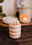 The Mimi & August Ceramic Match Pot, filled with cotton swabs, rests on a wooden surface beside a lit candle and chic plate—an ideal functional accessory to enhance your candle setup.