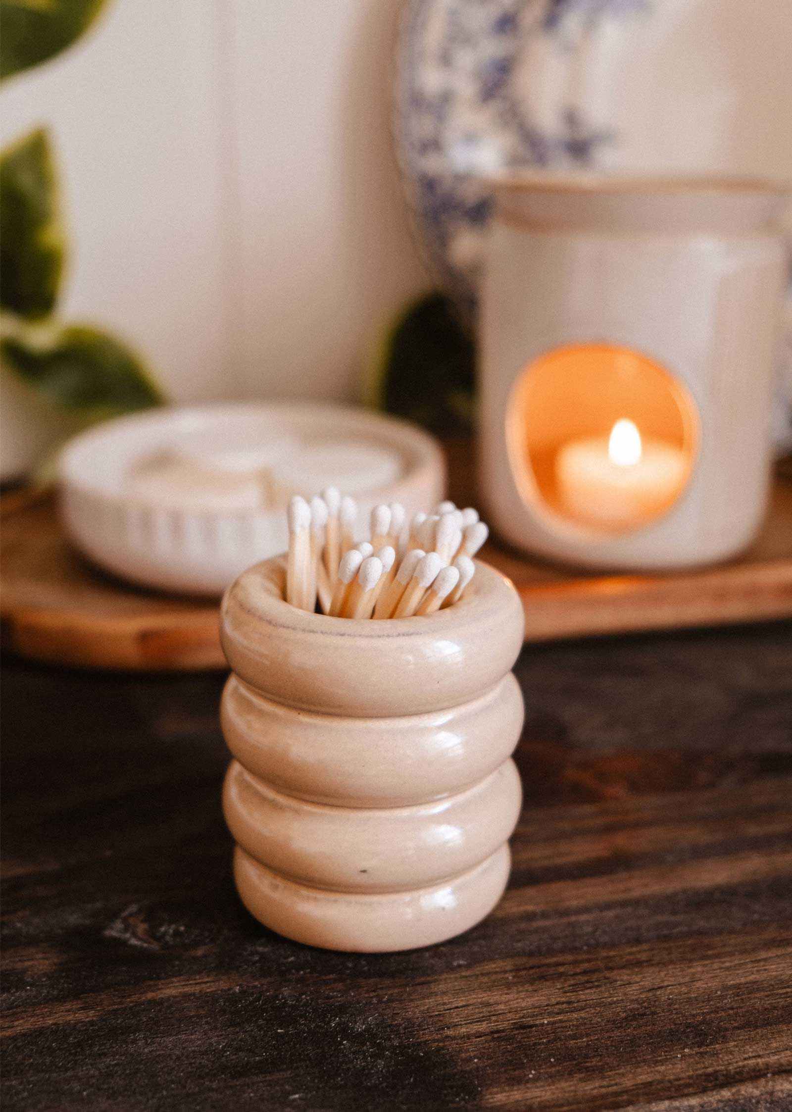 The Mimi & August Ceramic Match Pot, filled with cotton swabs, rests on a wooden surface beside a lit candle and chic plate—an ideal functional accessory to enhance your candle setup.