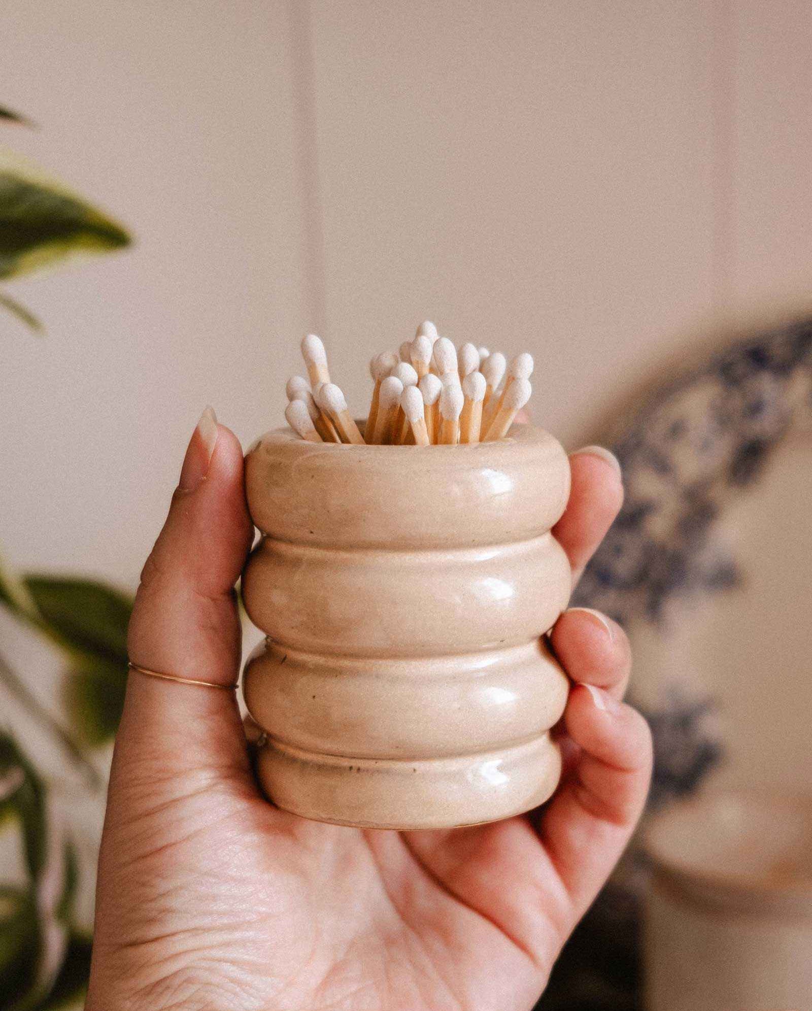 A hand holds the Mimi & August Ceramic Match Pot in beige, featuring a ribbed design and filled with cotton swabs. A blurred plant and plate appear in the background, highlighting the charm of this decorative candle accessory.
