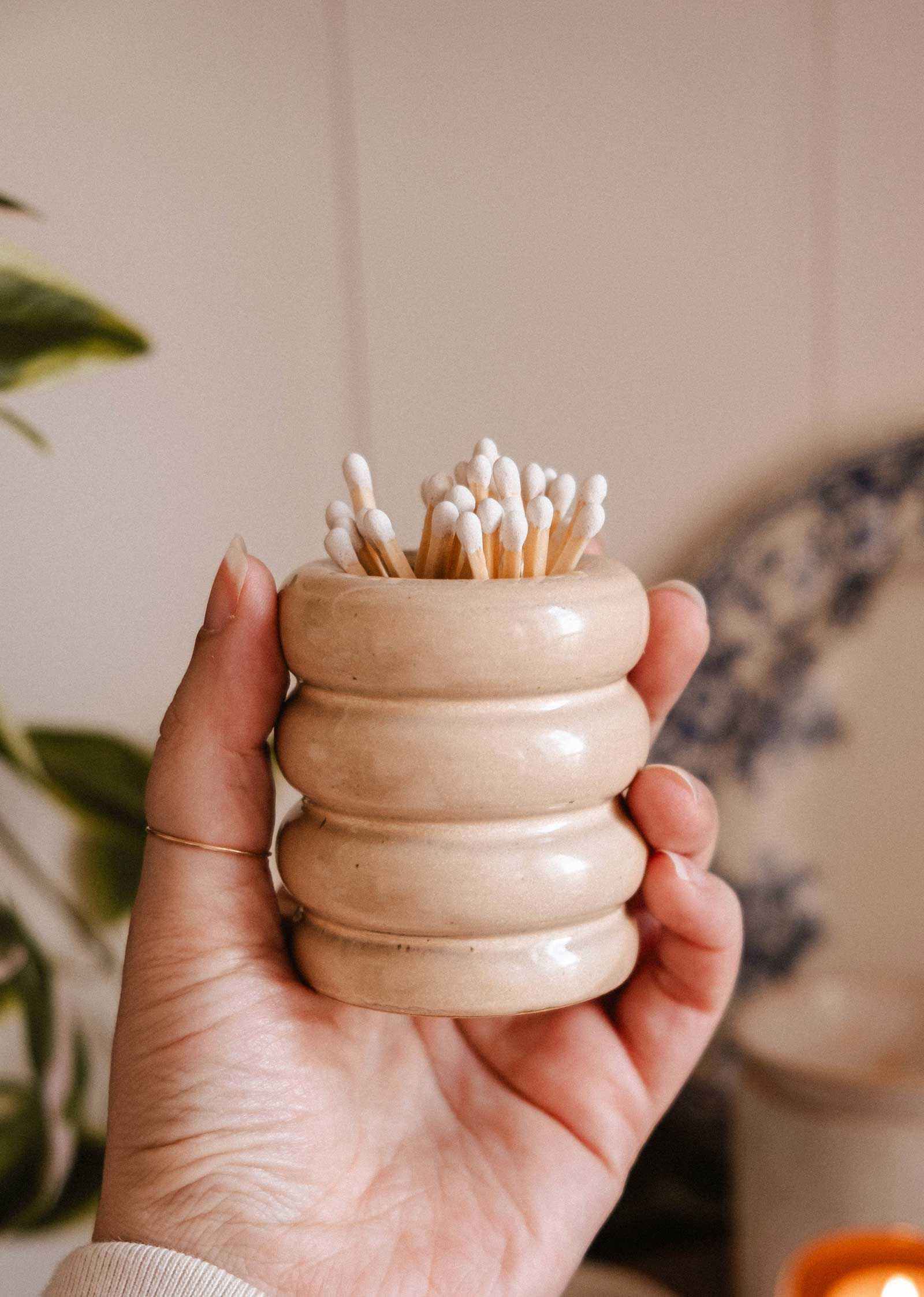 A hand holds the Mimi & August Ceramic Match Pot in beige, featuring a ribbed design and filled with cotton swabs. A blurred plant and plate appear in the background, highlighting the charm of this decorative candle accessory.