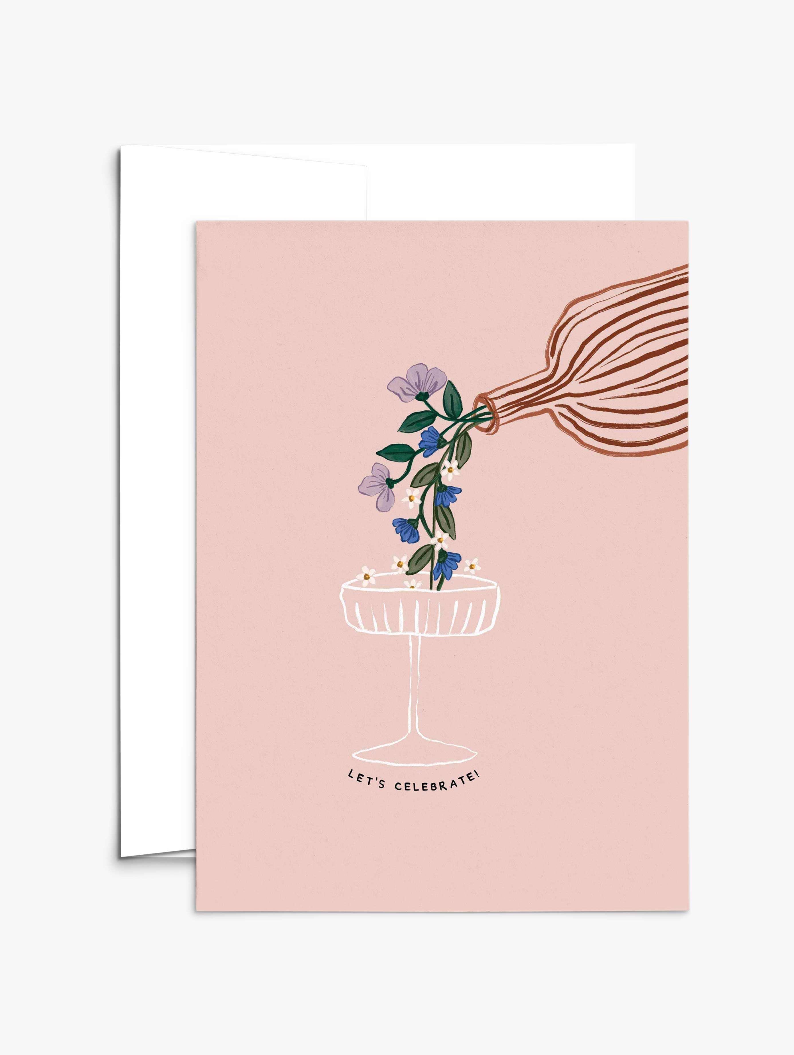 The Let's Celebrate Greeting Card by Mimi & August features a hand pouring flowers into a champagne glass on a pink background with "LET'S CELEBRATE!" below. Printed on recycled paper, it's the perfect card for any celebration.