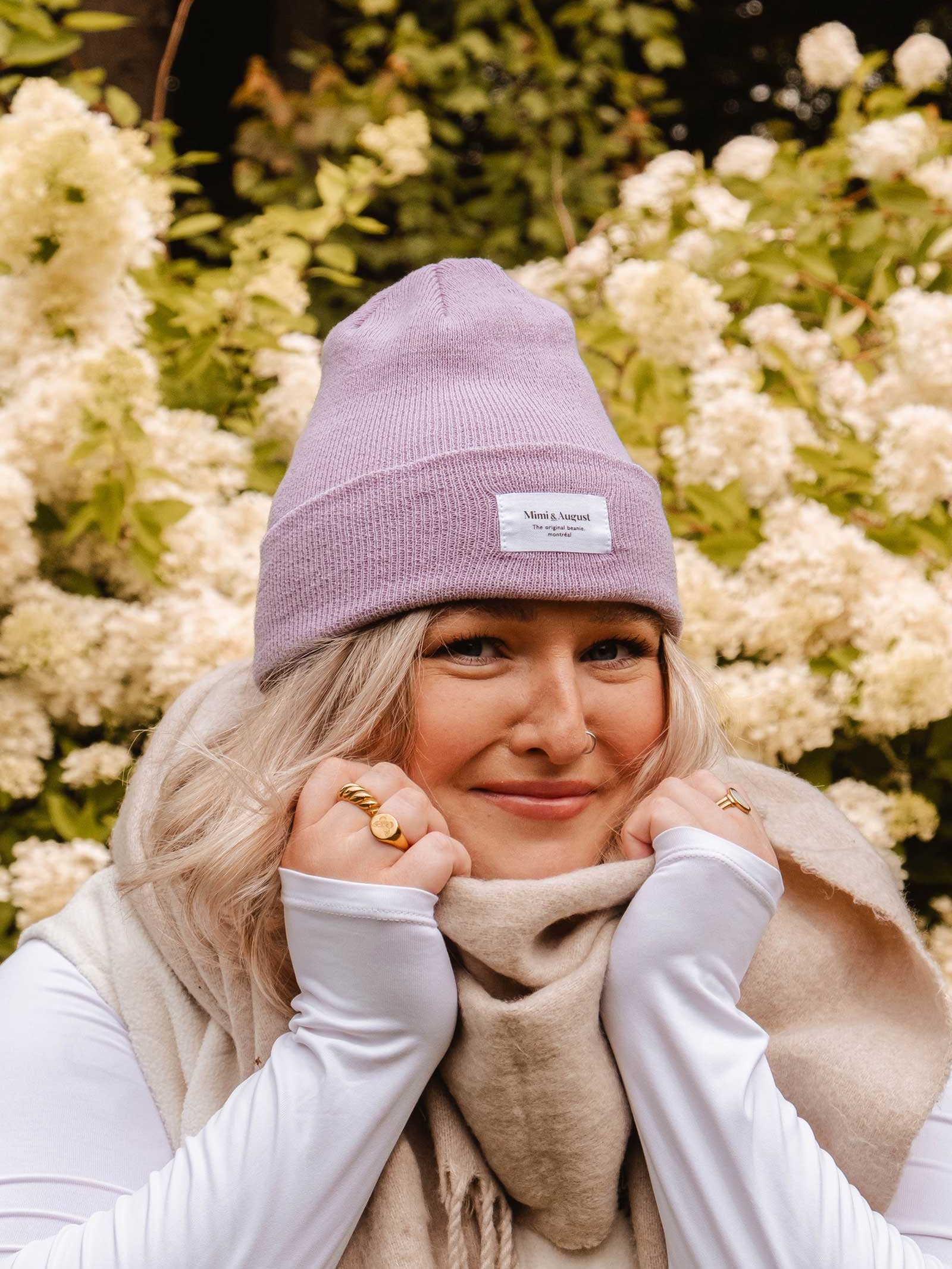 A person wearing a Mimi & August Lilac Beanie and a beige scarf smiles while holding their cozy accessory, standing in front of lush greenery and white flowers.