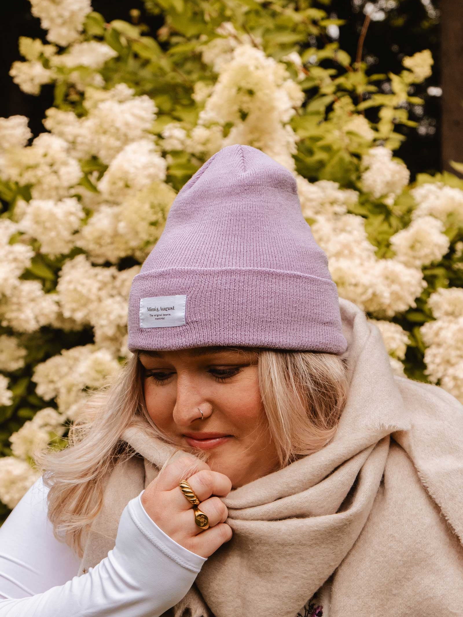 A person wearing a cozy Mimi & August Lilac Beanie and a beige scarf stands in front of white flowering bushes.