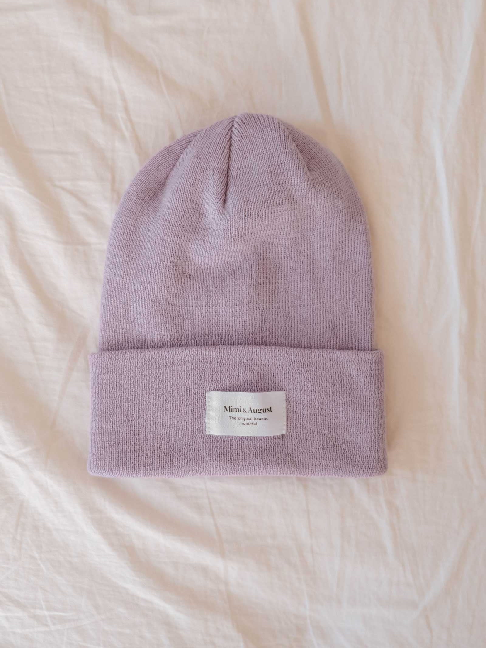 The Lilac Beanie by Mimi & August, a lilac knit with a folded edge and a rectangular white label on a beige fabric background, makes for the perfect cozy accessory. This unisex knit is both stylish and versatile.