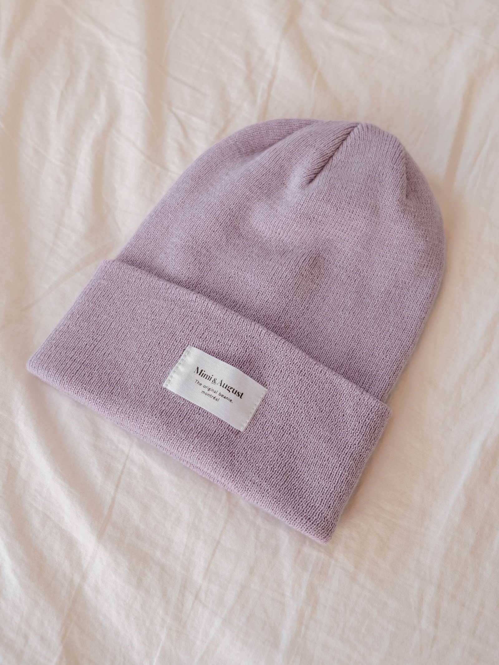 A cozy lilac beanie from Mimi & August featuring a white rectangular label, beautifully displayed on a soft beige fabric background, making it an ideal accessory for everyone.