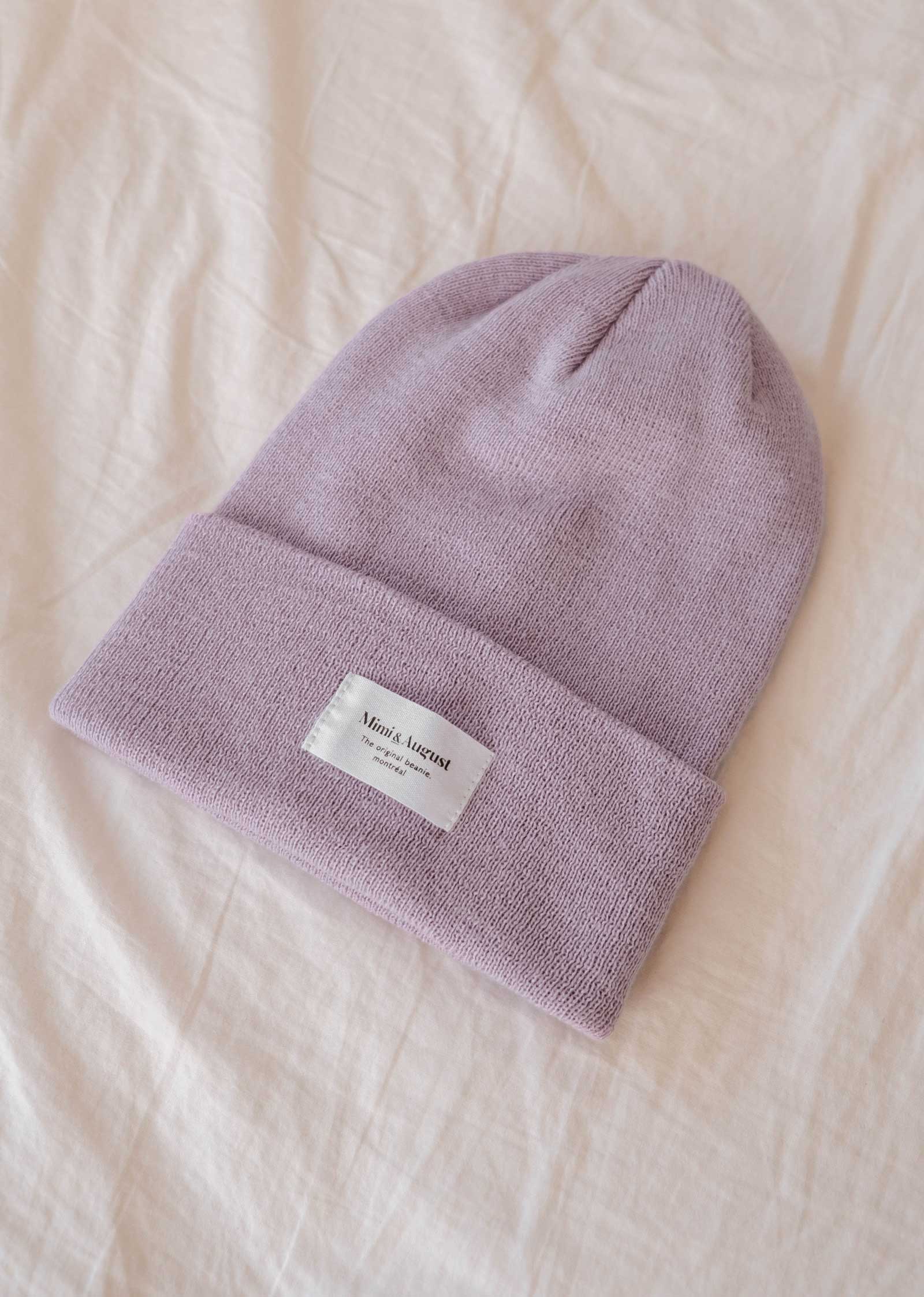 A cozy lilac beanie from Mimi & August featuring a white rectangular label, beautifully displayed on a soft beige fabric background, making it an ideal accessory for everyone.