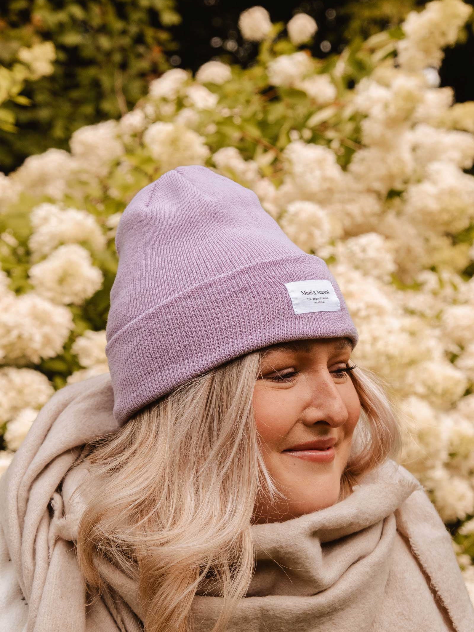 A person wearing a Lilac Beanie from Mimi & August and a cream unisex knit scarf is standing in front of white flowering shrubs.