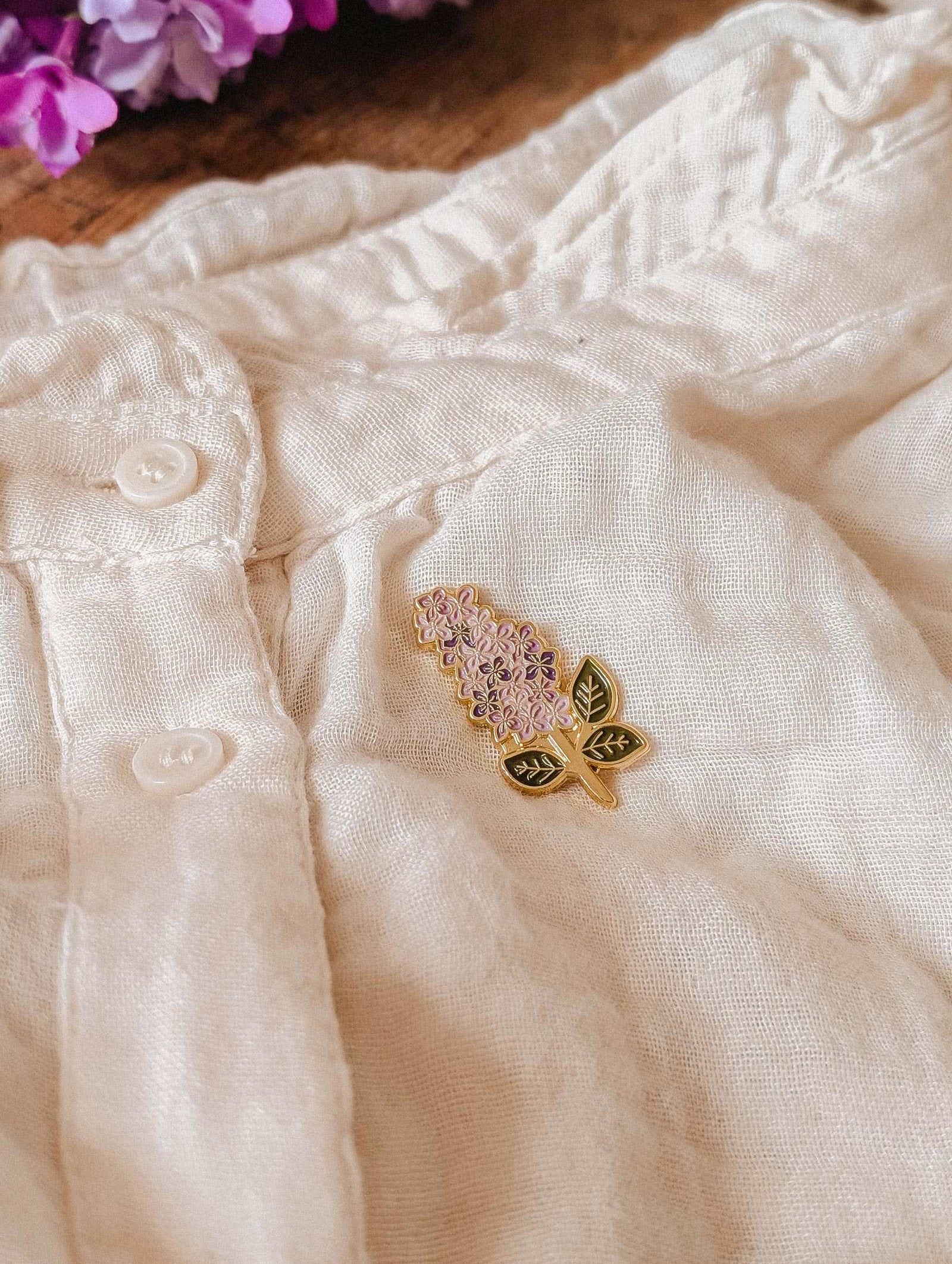The Mimi & August Lilac Enamel Pin in gold tone brass is attached to a cream linen garment with buttons, with lilac flowers visible in the upper left corner.