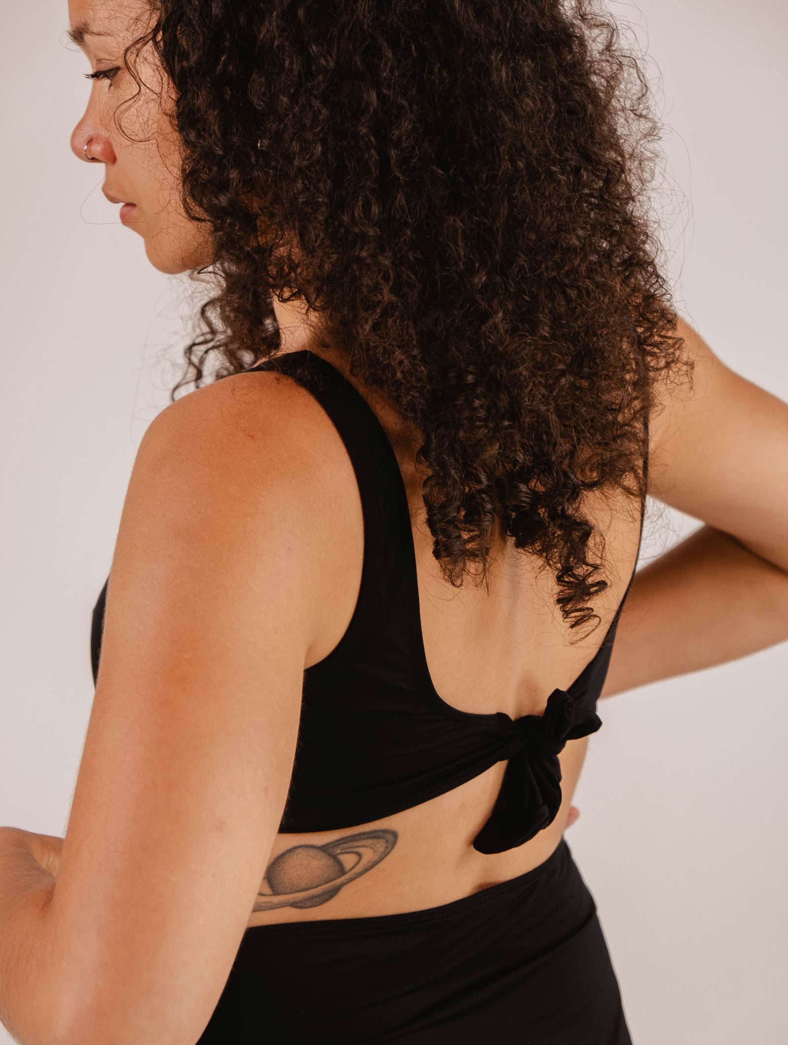 A woman with curly hair and a visible nose piercing is wearing a Mimi & August Lima Black Bralette Bikini Top with an adjustable back tied in a bow, exposing a tattoo of Saturn on her left side. She is looking to the left.