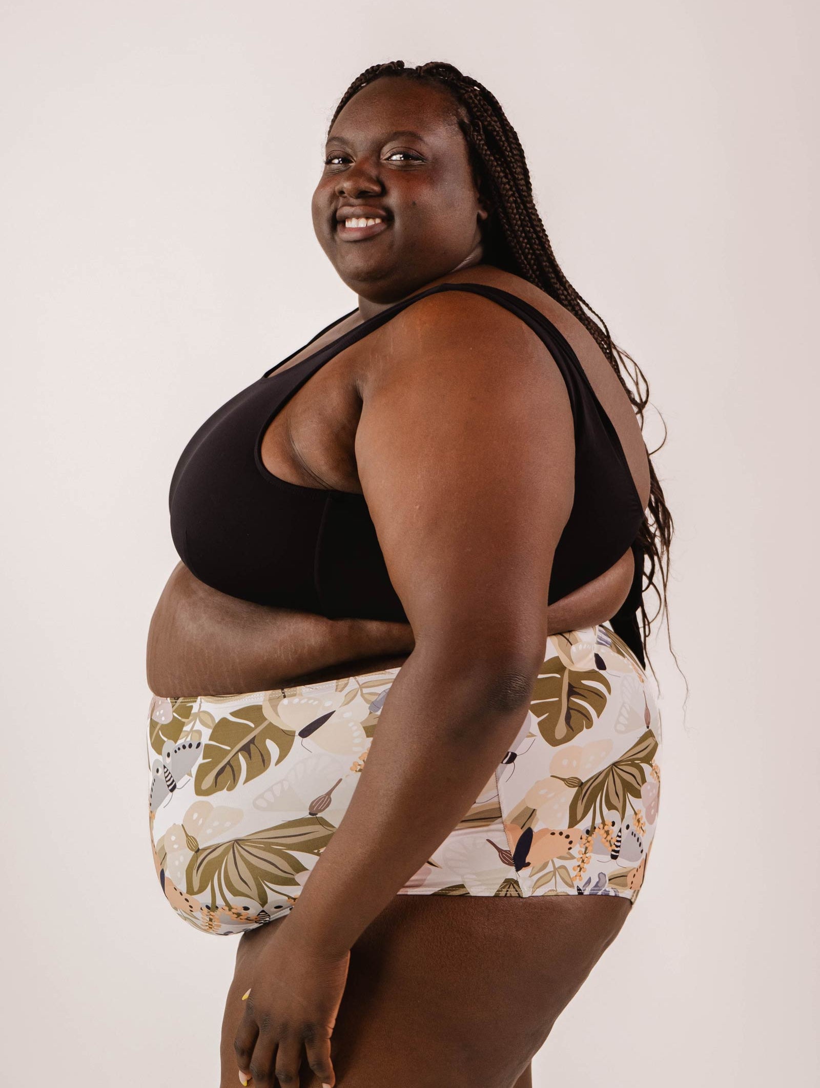 A person smiling and posing in a Mimi & August Lima Black Bralette Bikini Top with adjustable back and patterned shorts against a white background.