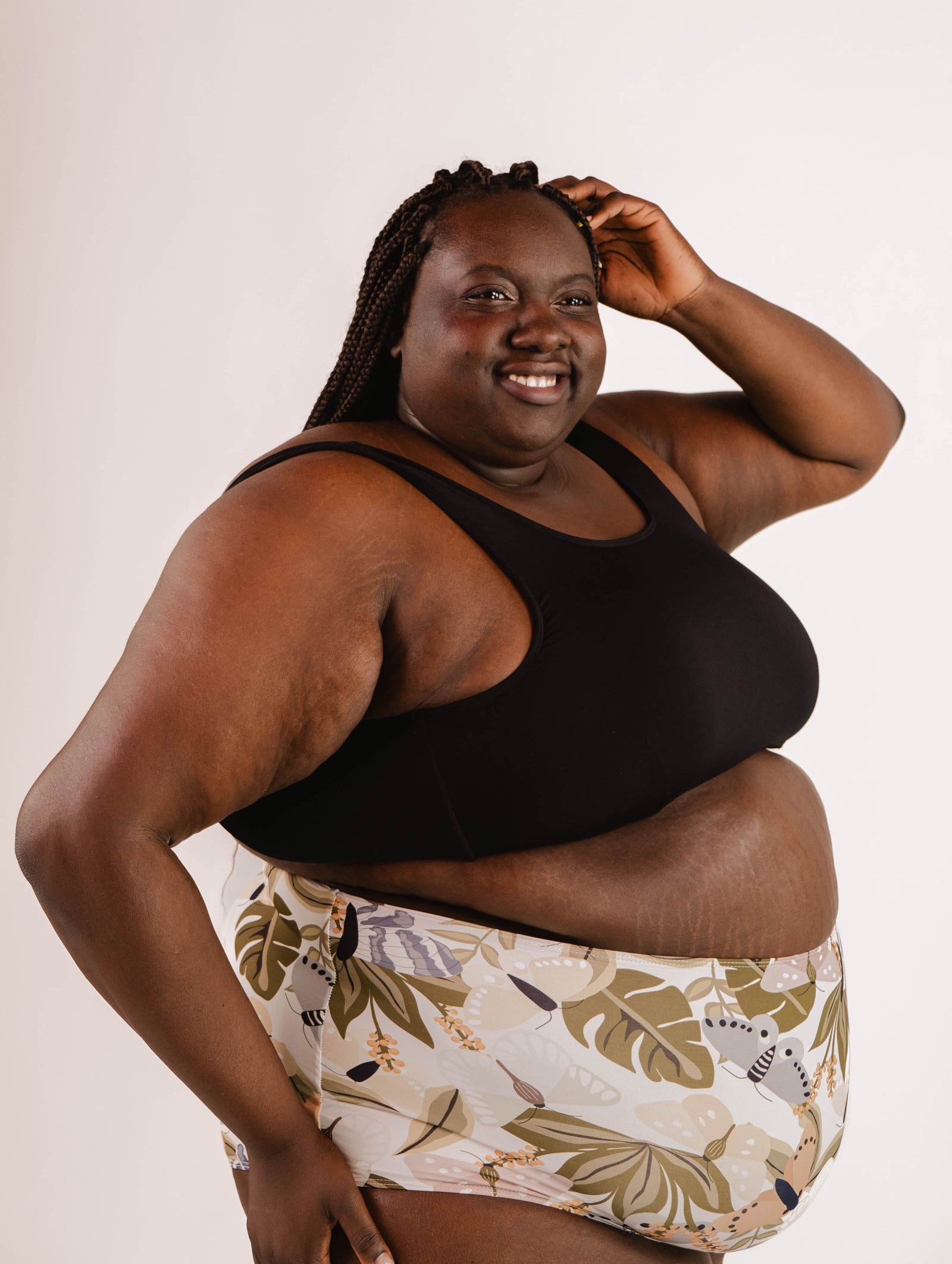 A person with braided hair smiles while posing in a Lima Black Bralette Bikini Top by Mimi & August, featuring adjustable back and removable pads, paired with patterned shorts against a plain background.