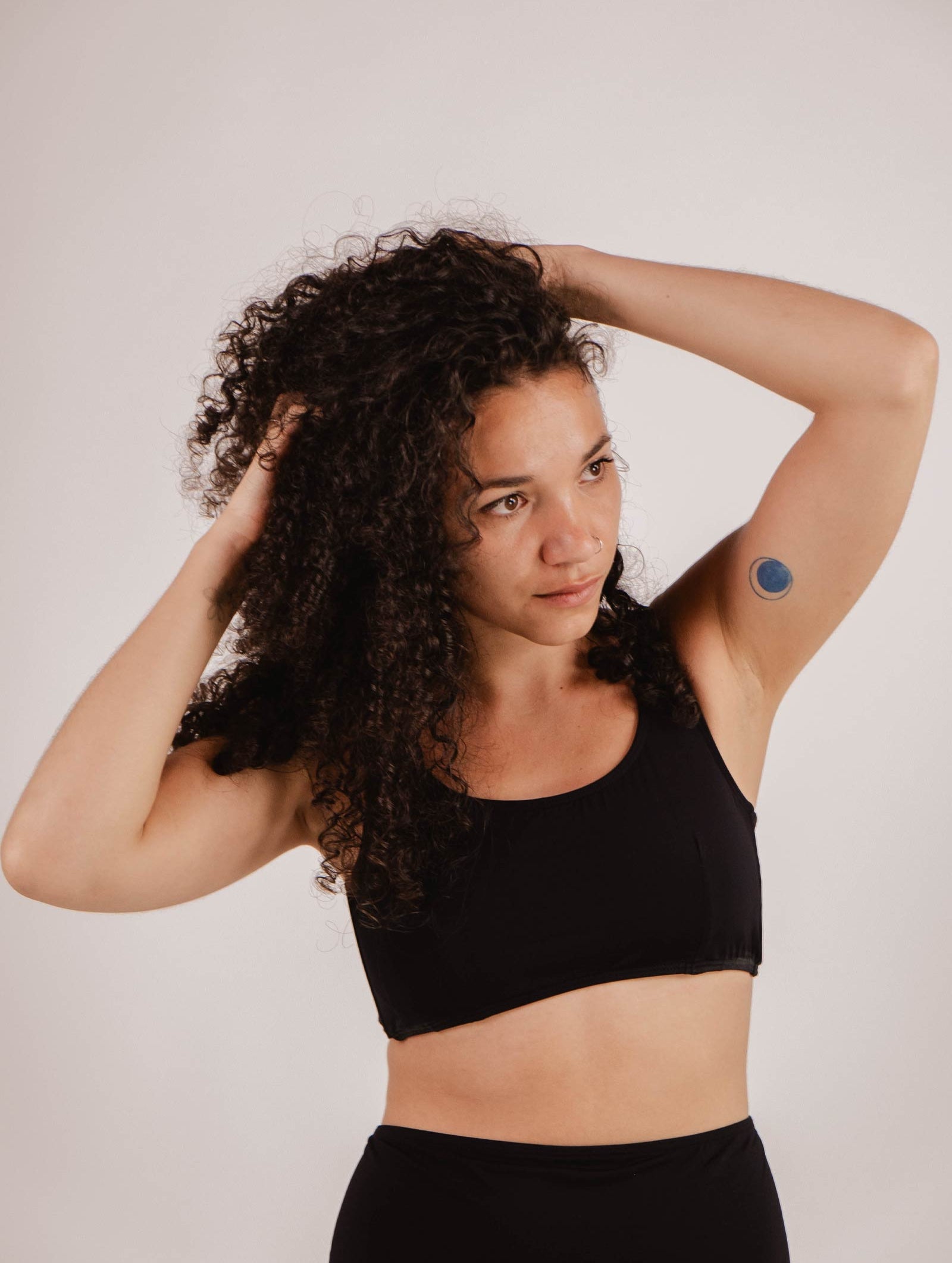 A person with curly hair wearing a Mimi & August Lima Black Bralette Bikini Top and black bottoms touches their hair. There is a small blue circular patch on the upper part of their left arm.