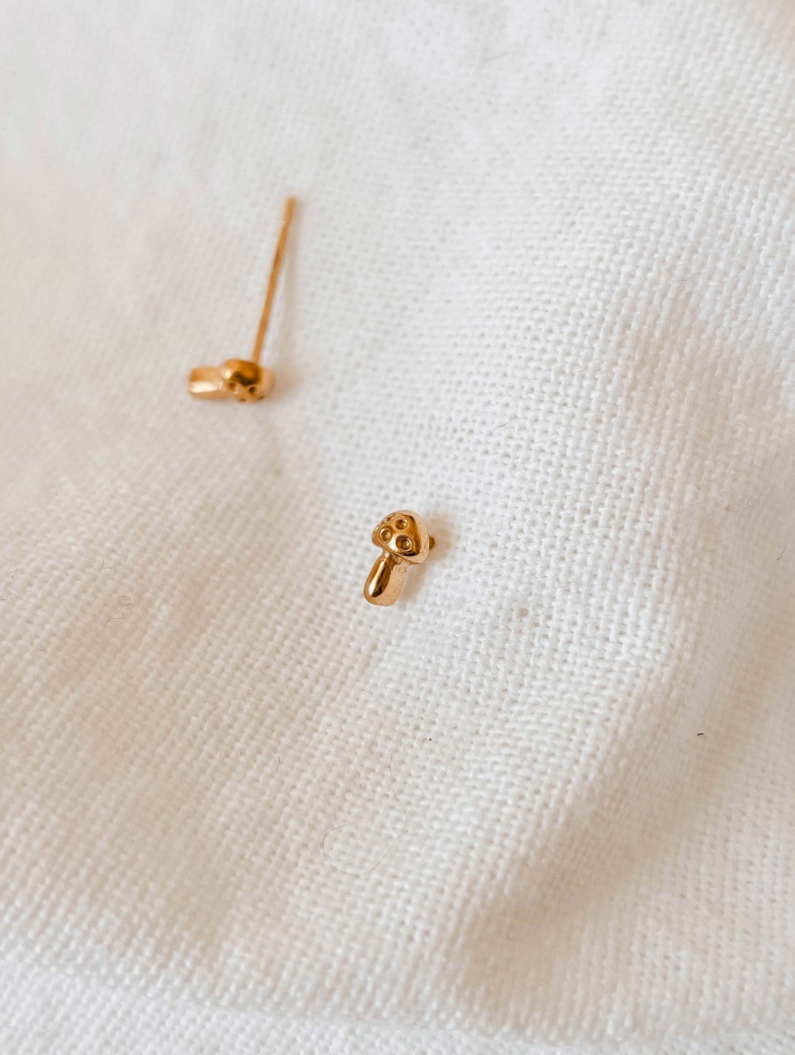 These charming gold-plated earrings, crafted by Mimi & August and named "Little Mushroom," elegantly rest on a white fabric surface, showcasing their unique handmade-in-USA quality.