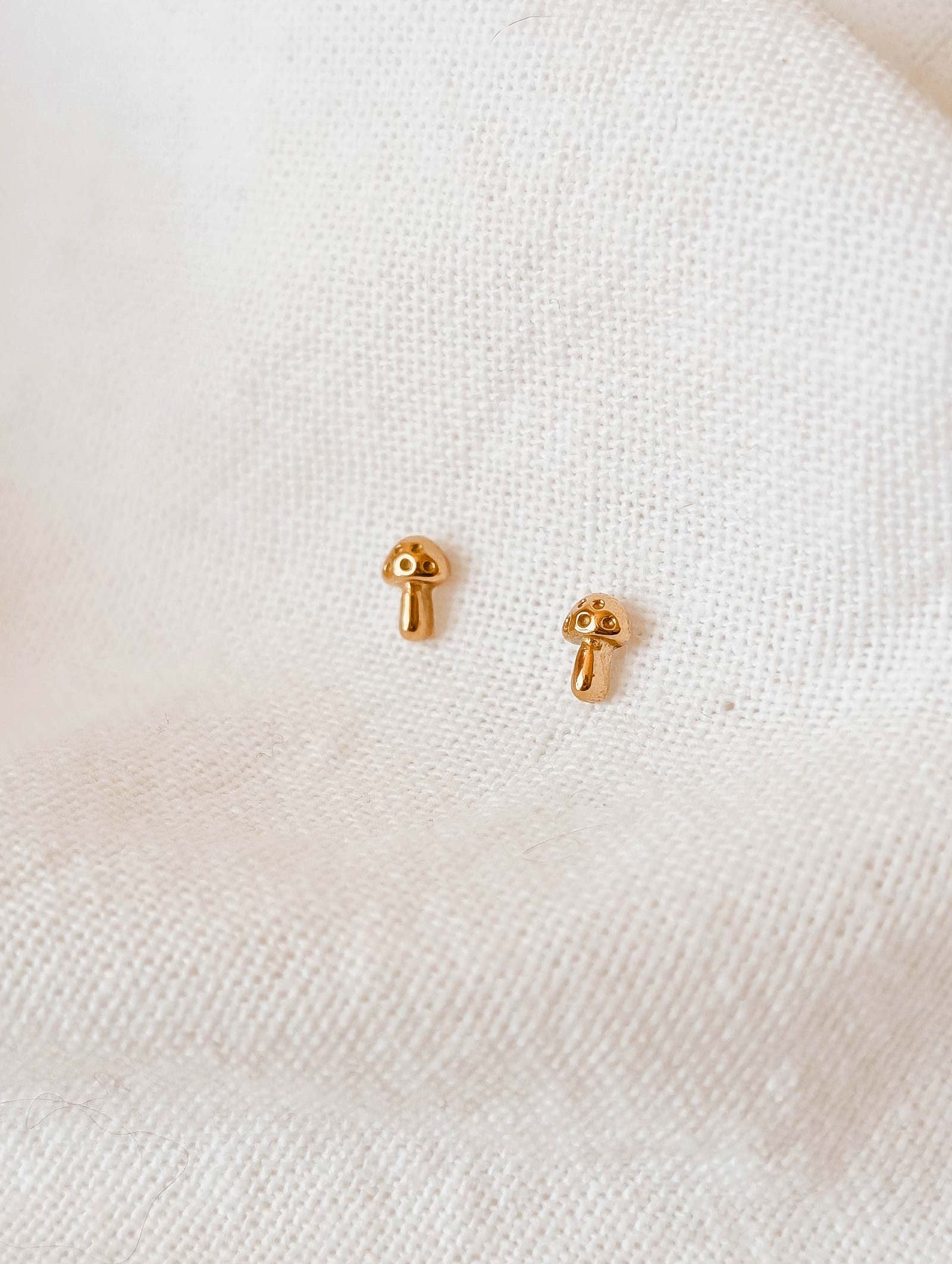 Mimi & August's Little Mushroom Gold Plated Earrings, charmingly handmade in the USA, glisten delicately against a textured white fabric backdrop.