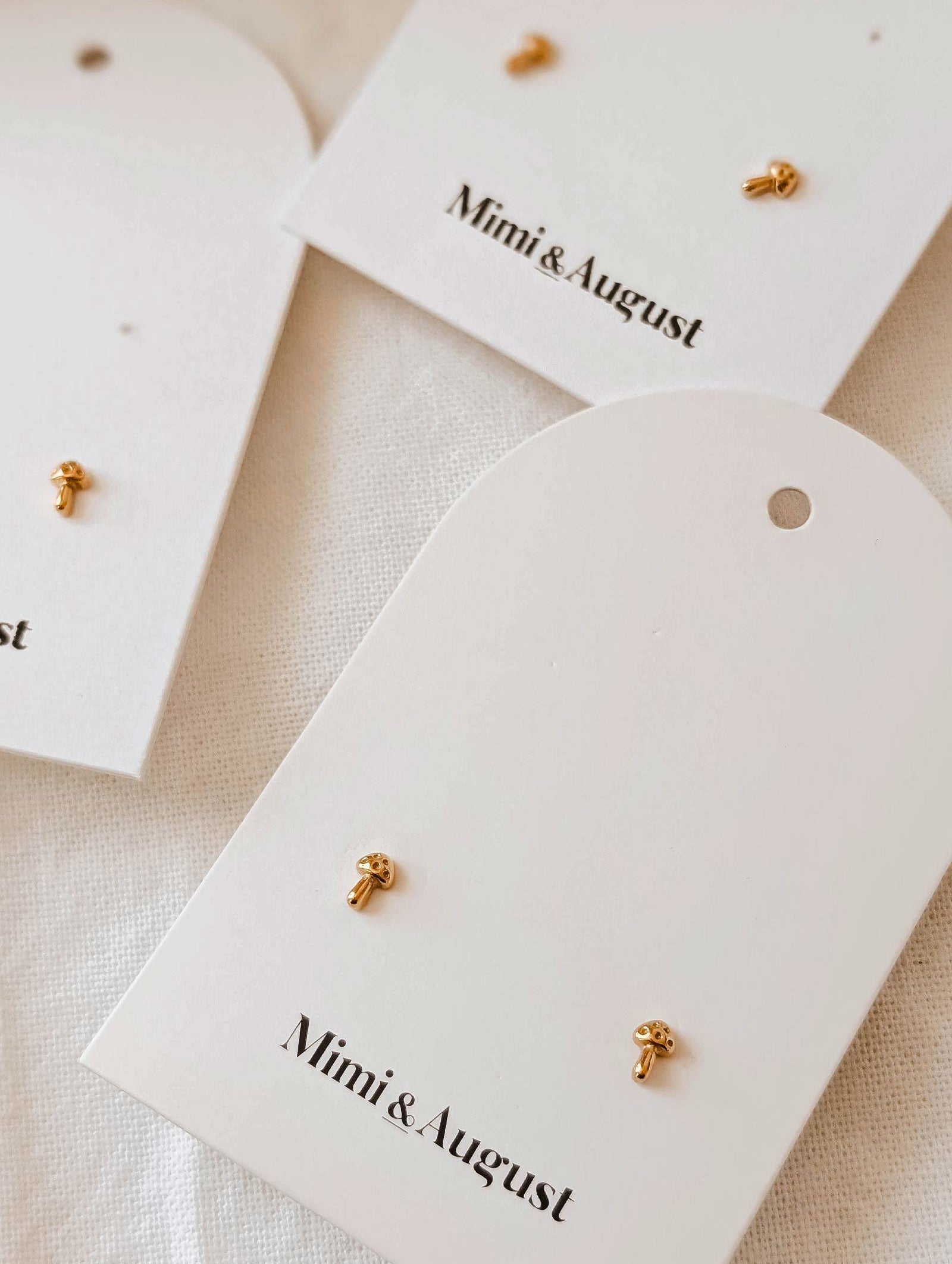 Three pairs of gold-plated Little Mushroom earrings by Mimi & August are elegantly displayed on white cards.