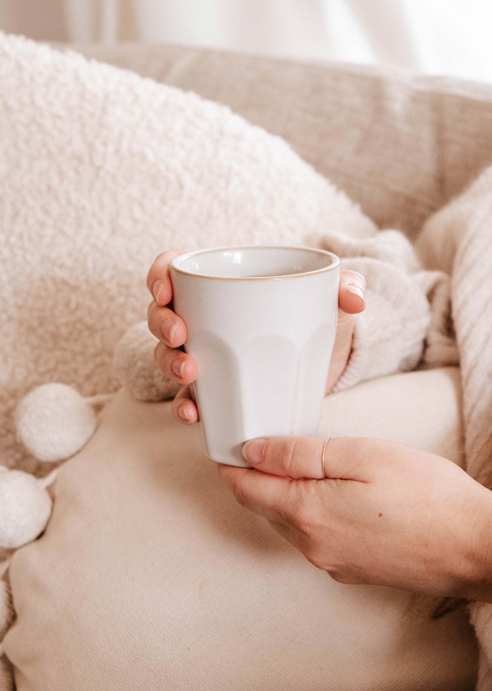 A person holds a white ceramic mug with both hands on a beige couch with a textured blanket and a linen tea towel nearby—ideal for gifting the Mimi & August Little Moments - Gift Box.