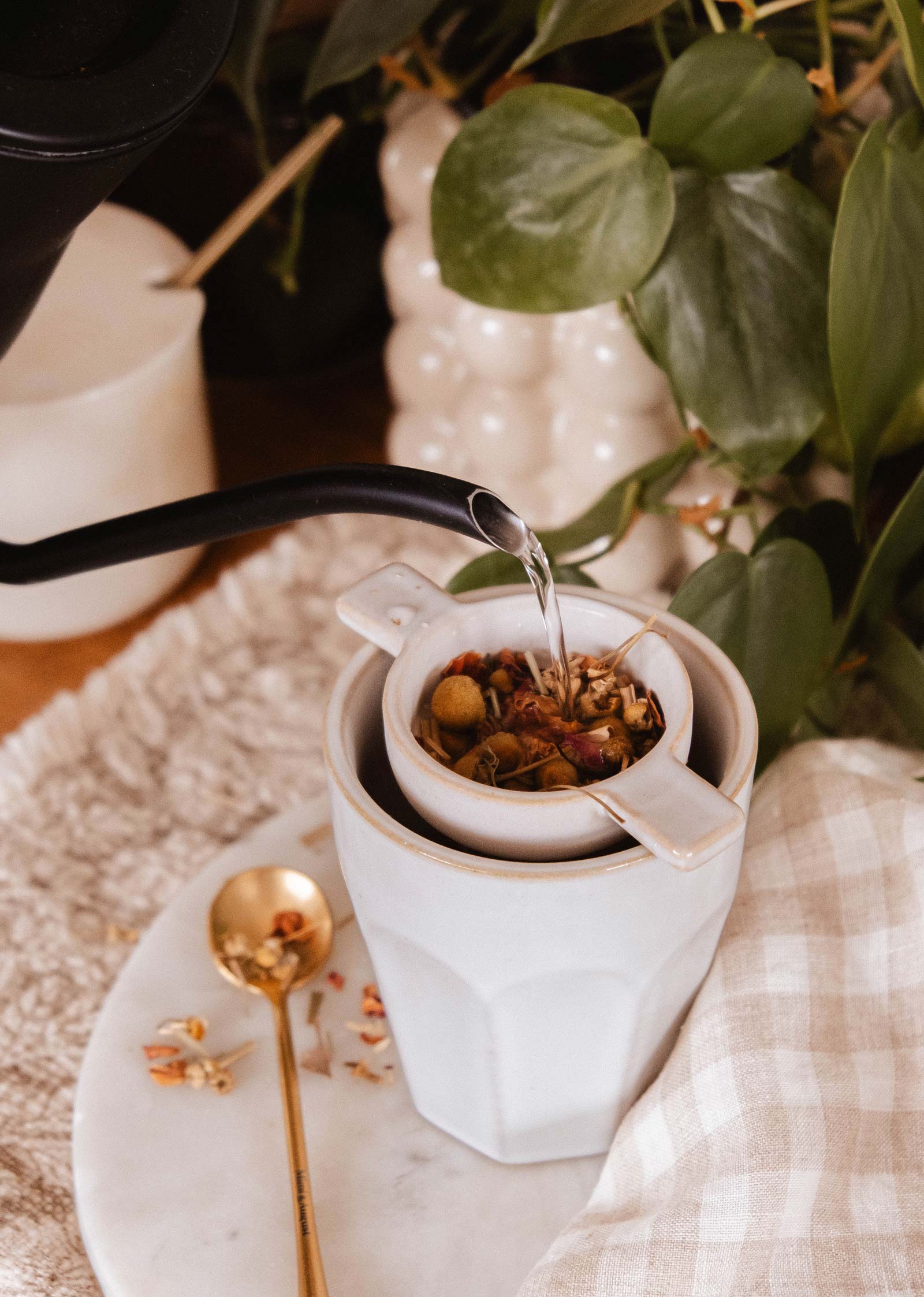 Hot water from a black kettle is poured over loose leaf tea in a ceramic strainer atop a white mug, with a gold spoon, plant, and linen towel—creating a cozy scene from the Mimi & August Little Moments - Gift Box.