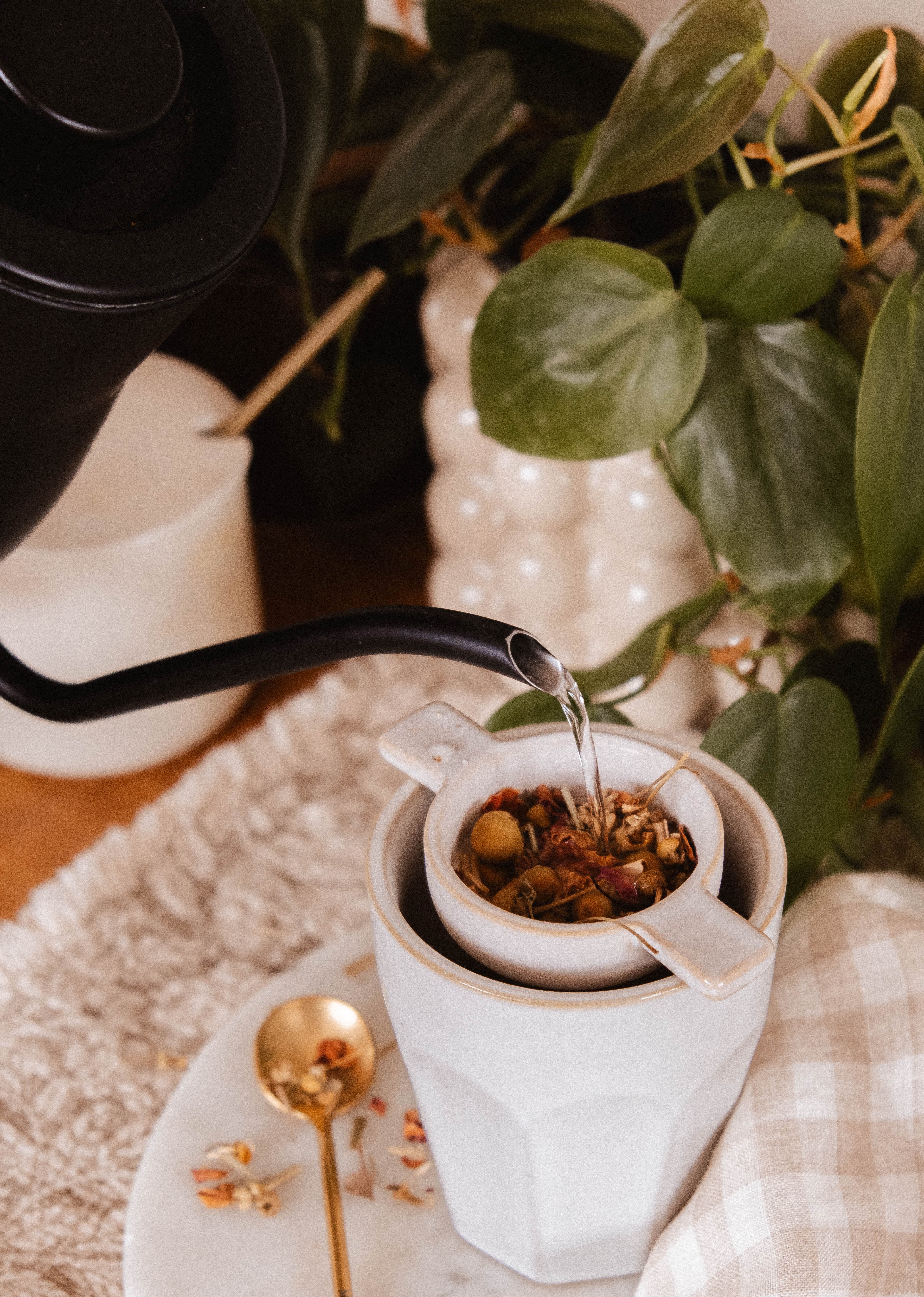 Hot water pours through Mimi & August's The Infusion Nest – Ceramic Tea Strainer, filling a handcrafted white cup. A gold spoon and a green plant complete this serene tea moment.