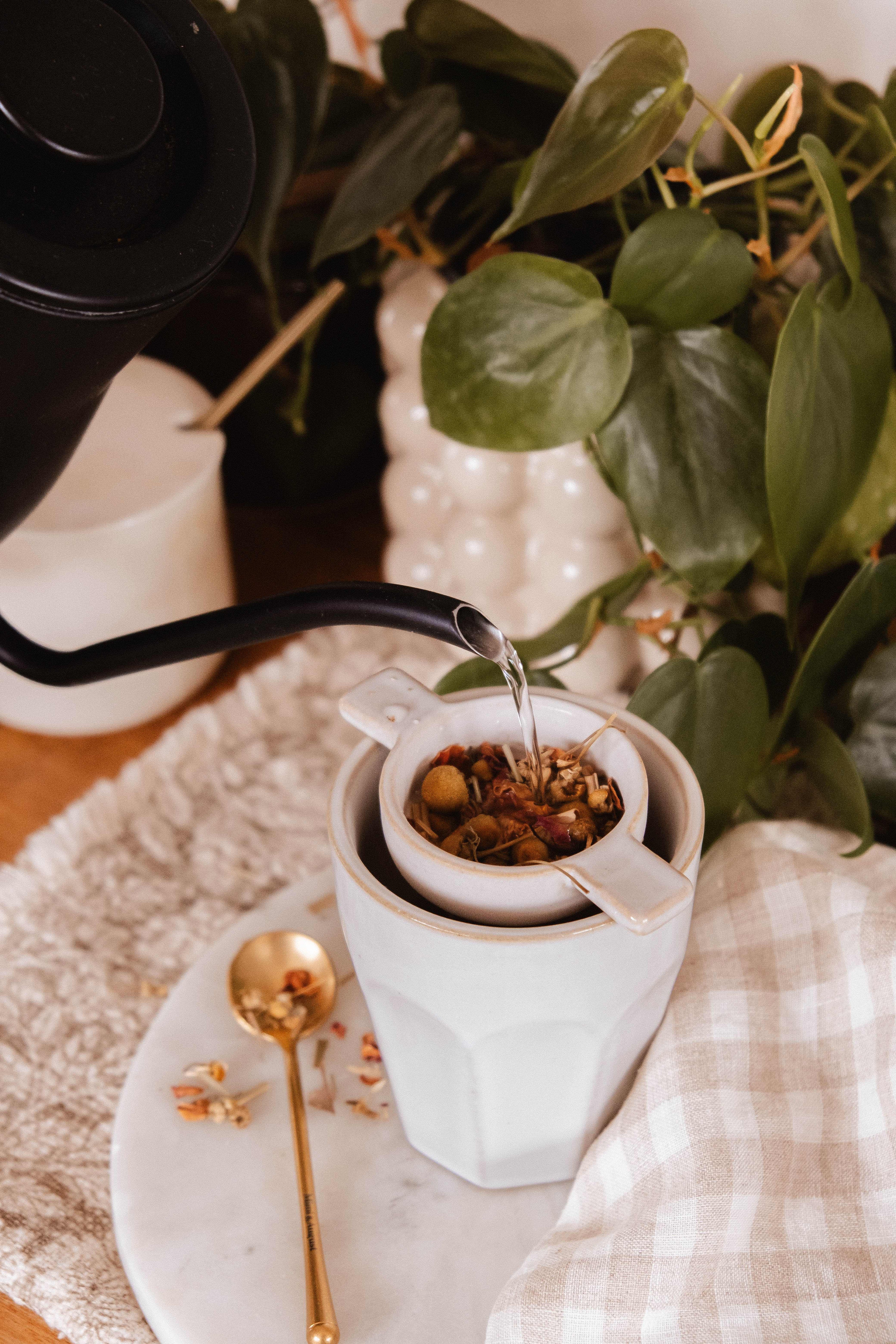 Hot water pours through Mimi & August's The Infusion Nest – Ceramic Tea Strainer, filling a handcrafted white cup. A gold spoon and a green plant complete this serene tea moment.