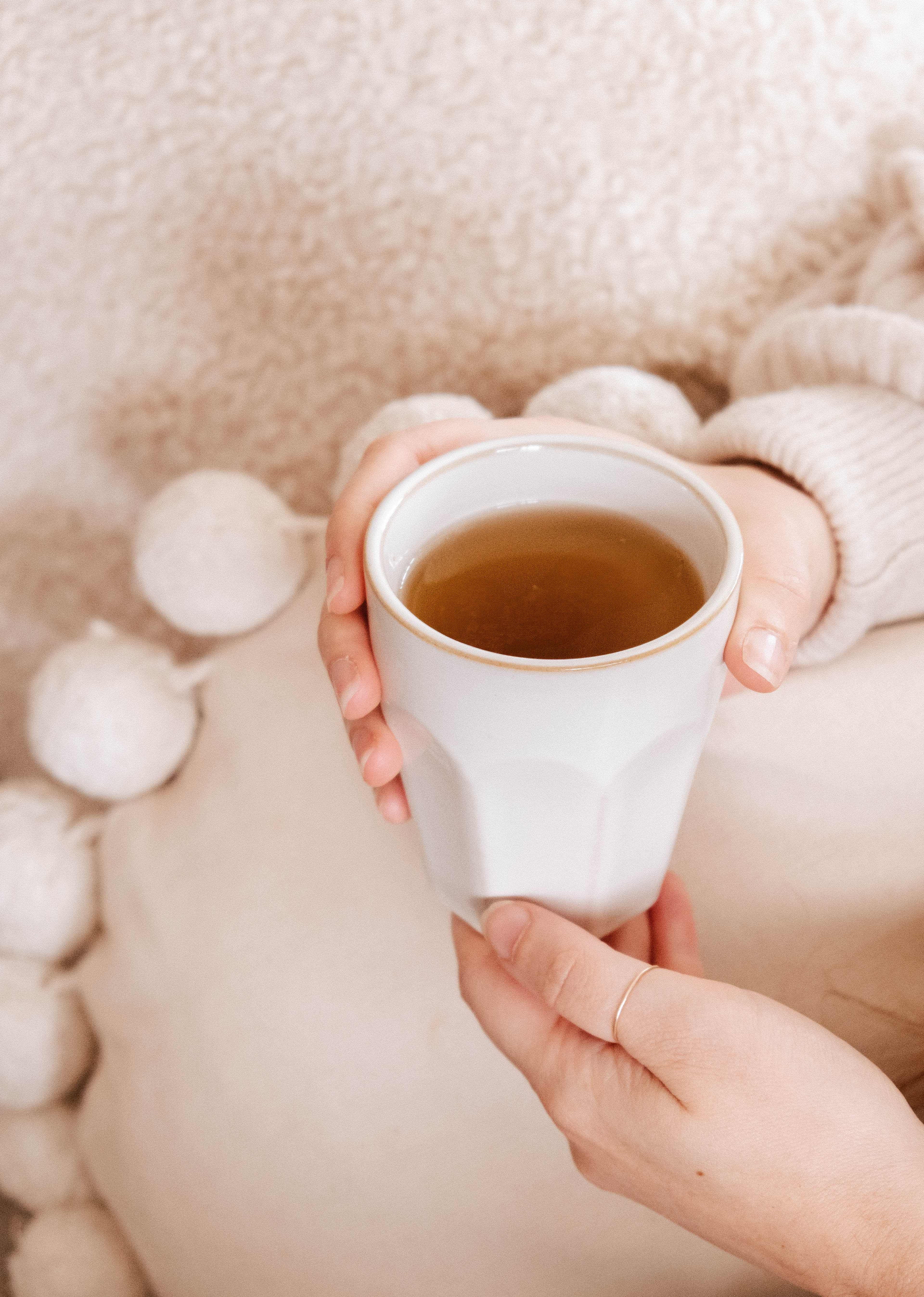 A person in a light sweater holds the Soft Morning – Ceramic Cup by Mimi & August, filled with tea, while sitting against a textured pillow with pom-pom accents. The handcrafted cup is microwave and dishwasher safe.