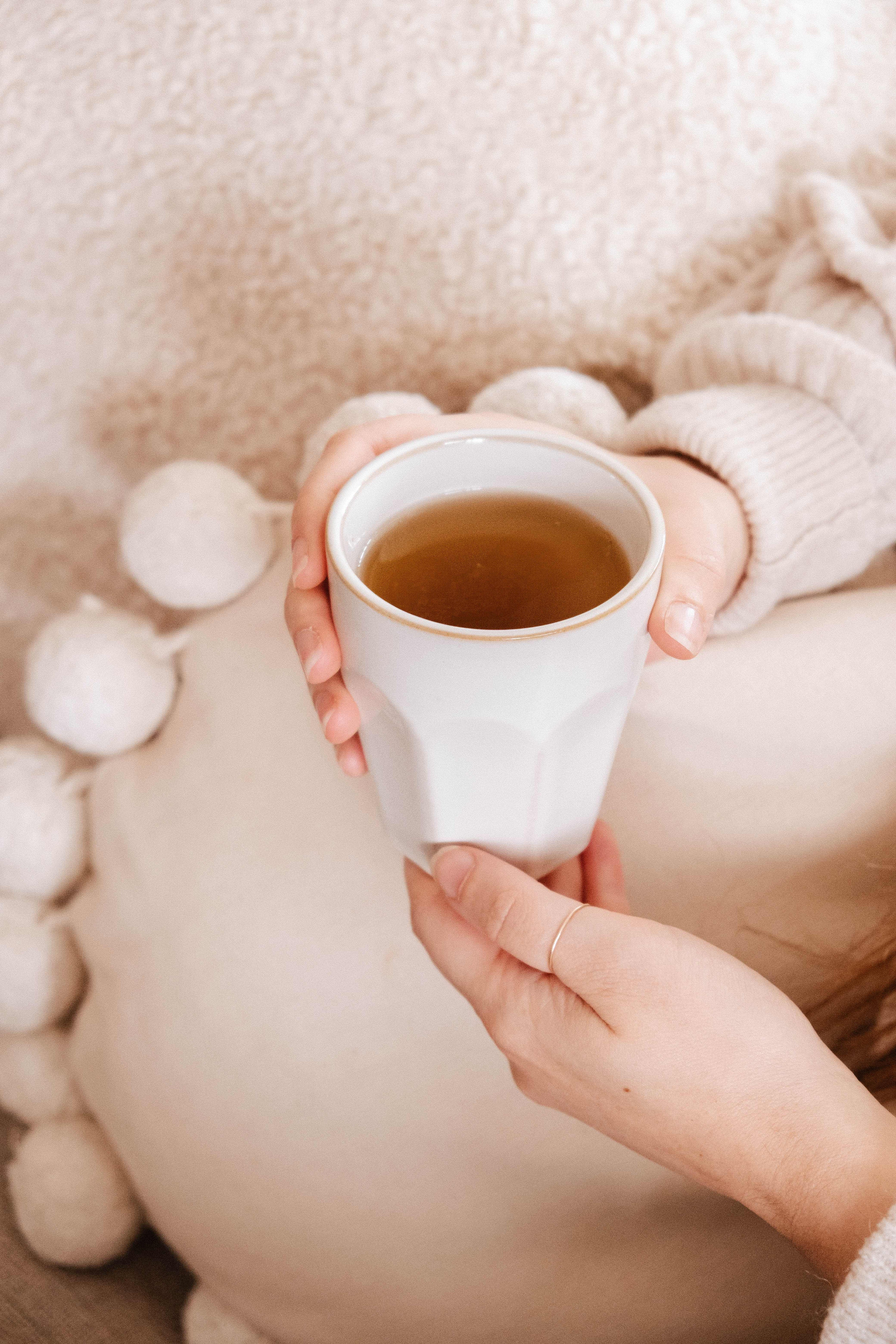 A person in a light sweater holds the Soft Morning – Ceramic Cup by Mimi & August, filled with tea, while sitting against a textured pillow with pom-pom accents. The handcrafted cup is microwave and dishwasher safe.