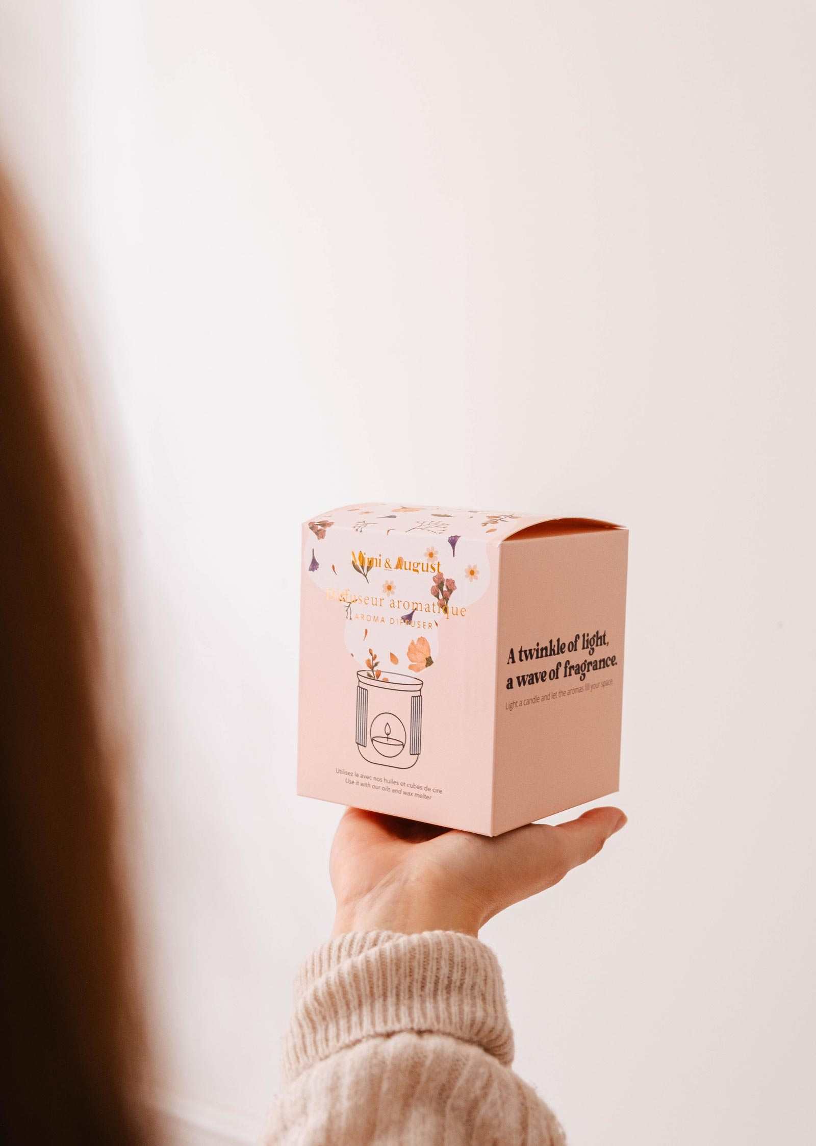 A person holds a decorative box featuring floral designs and fragrance text, along with the Lueur – Aromatic Ceramic Diffuser by Mimi & August, set against a neutral background.