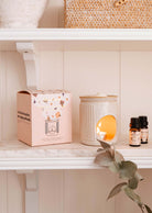 The Lueur – Aromatic Ceramic Diffuser by Mimi & August, with a lit candle inside, sits on a shelf next to scented oils and a decorative box, adorned with nearby eucalyptus leaves.