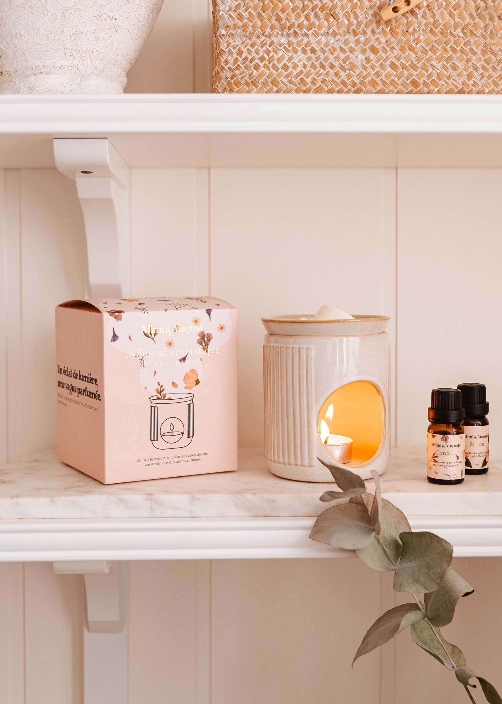 The Lueur – Aromatic Ceramic Diffuser by Mimi & August, with a lit candle inside, sits on a shelf next to scented oils and a decorative box, adorned with nearby eucalyptus leaves.