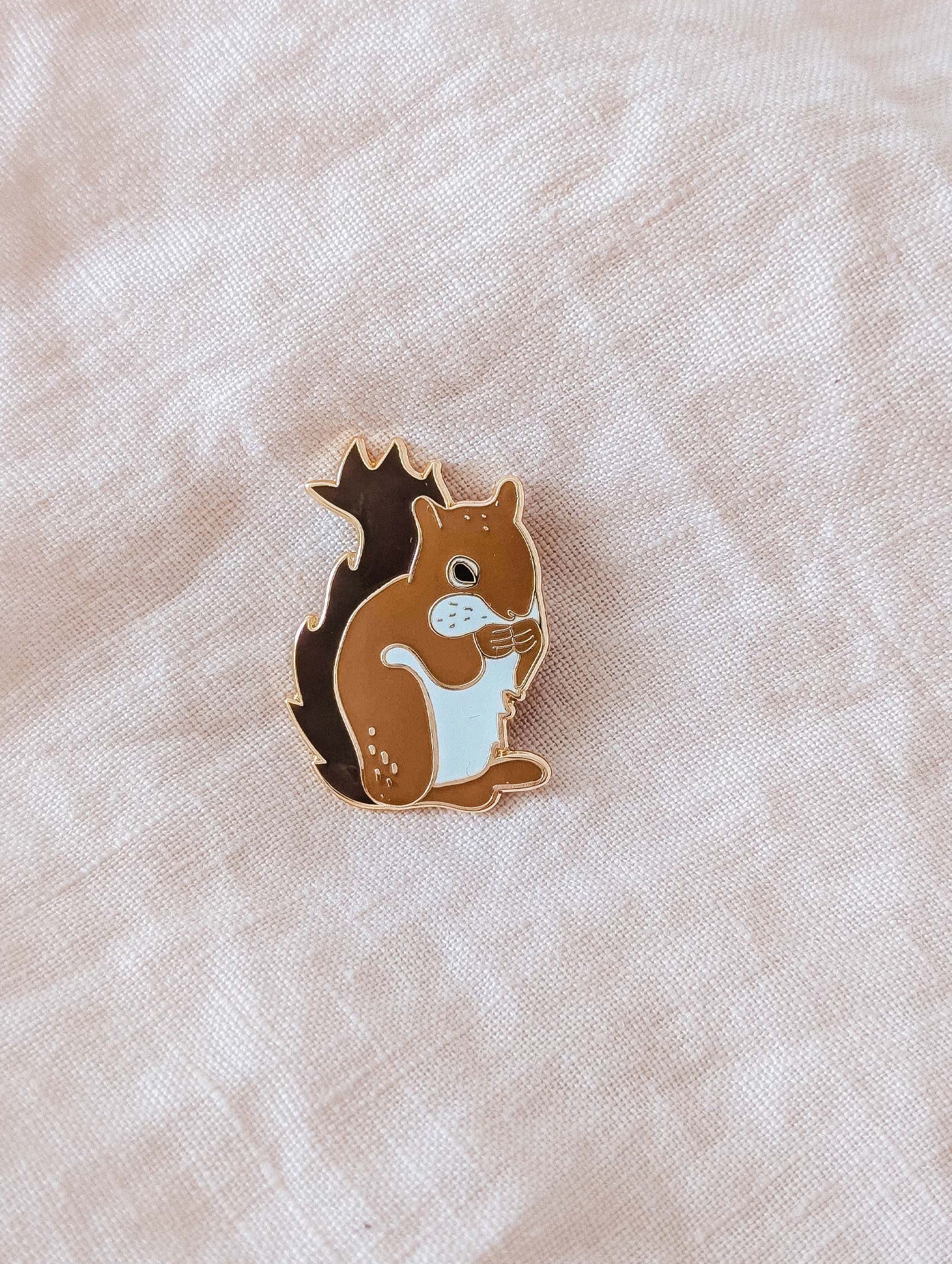 The Mimi & August MTL Squirrel Enamel Pin, in brown and white, rests on a light beige fabric—an adorable Montreal-inspired accessory ideal for animal lovers.