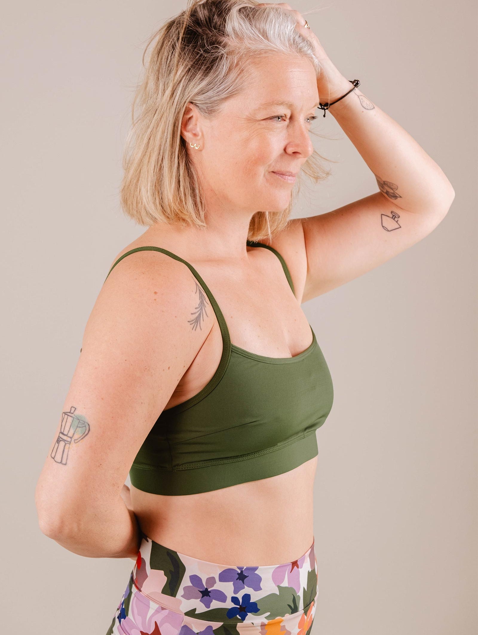 A mature woman with blonde hair in a Mimi & August Mango Amazonia Bralette Bikini Top and colorful leggings, posing with her hand on her head against a beige background.