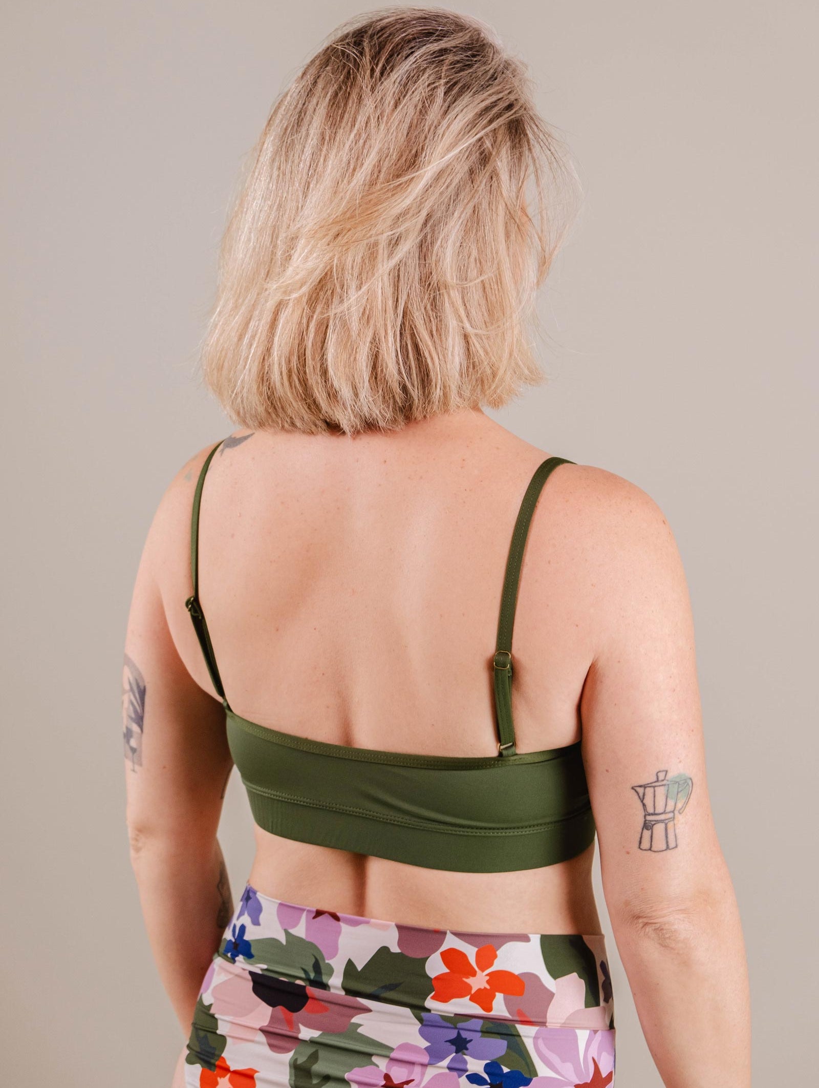 Woman with blonde bob haircut, wearing a green Mimi & August Mango Amazonia Bralette Bikini Top and floral leggings, viewed from the back. She has visible tattoos on her arms and upper back.