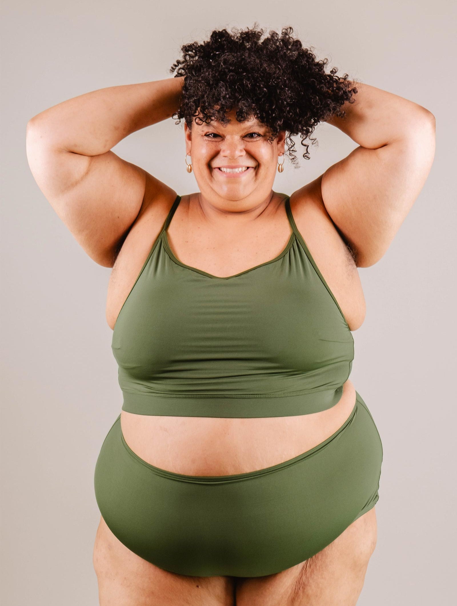 A confident woman in a Mimi & August Mango Amazonia Bralette Bikini Top and leggings, smiling as she poses with her hands lifting her curly hair.