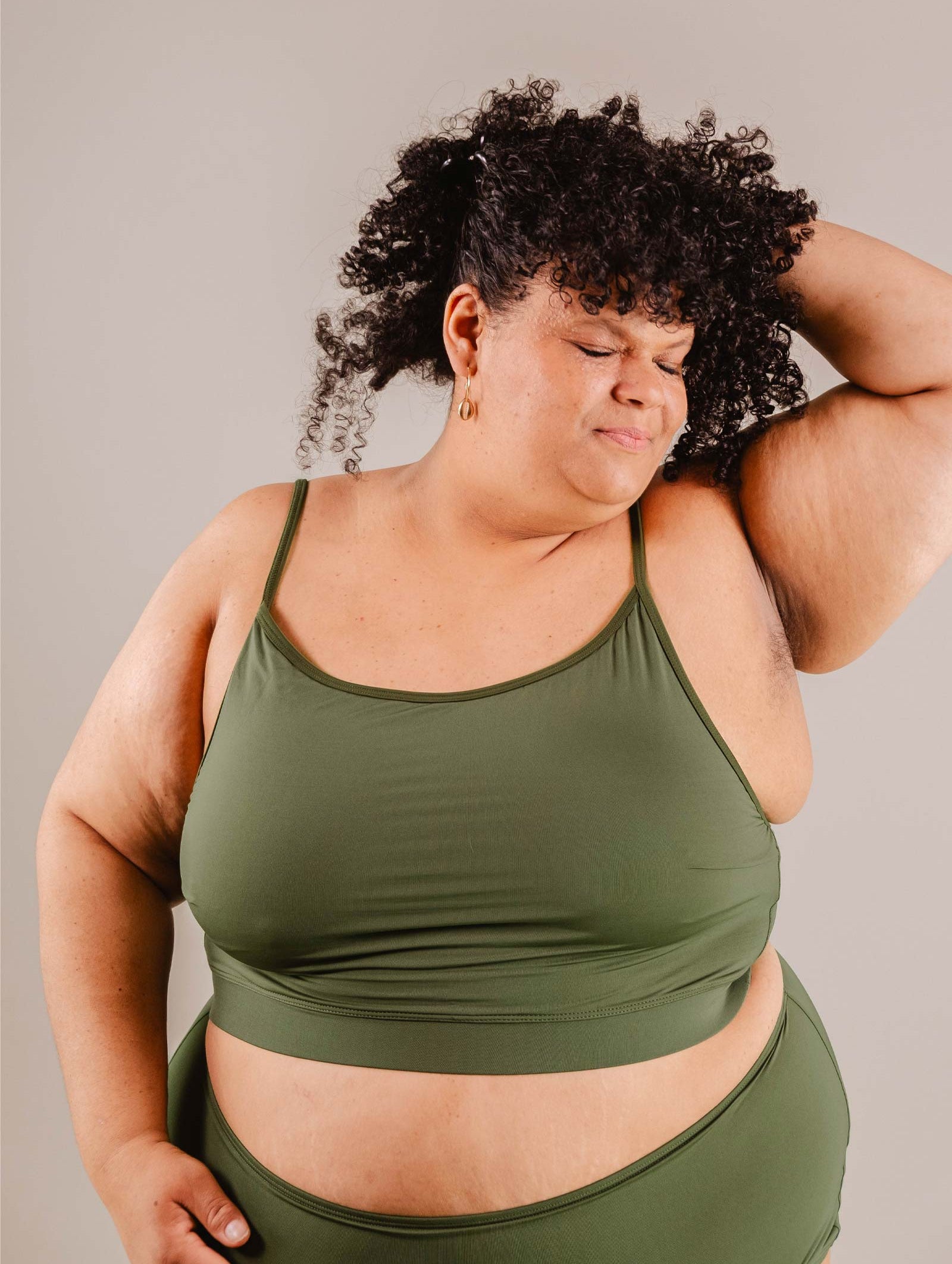 A plus-size woman in a Mimi & August Mango Amazonia Bralette Bikini Top posing with her hand on her head, her eyes closed and a slight smile, against a neutral background.