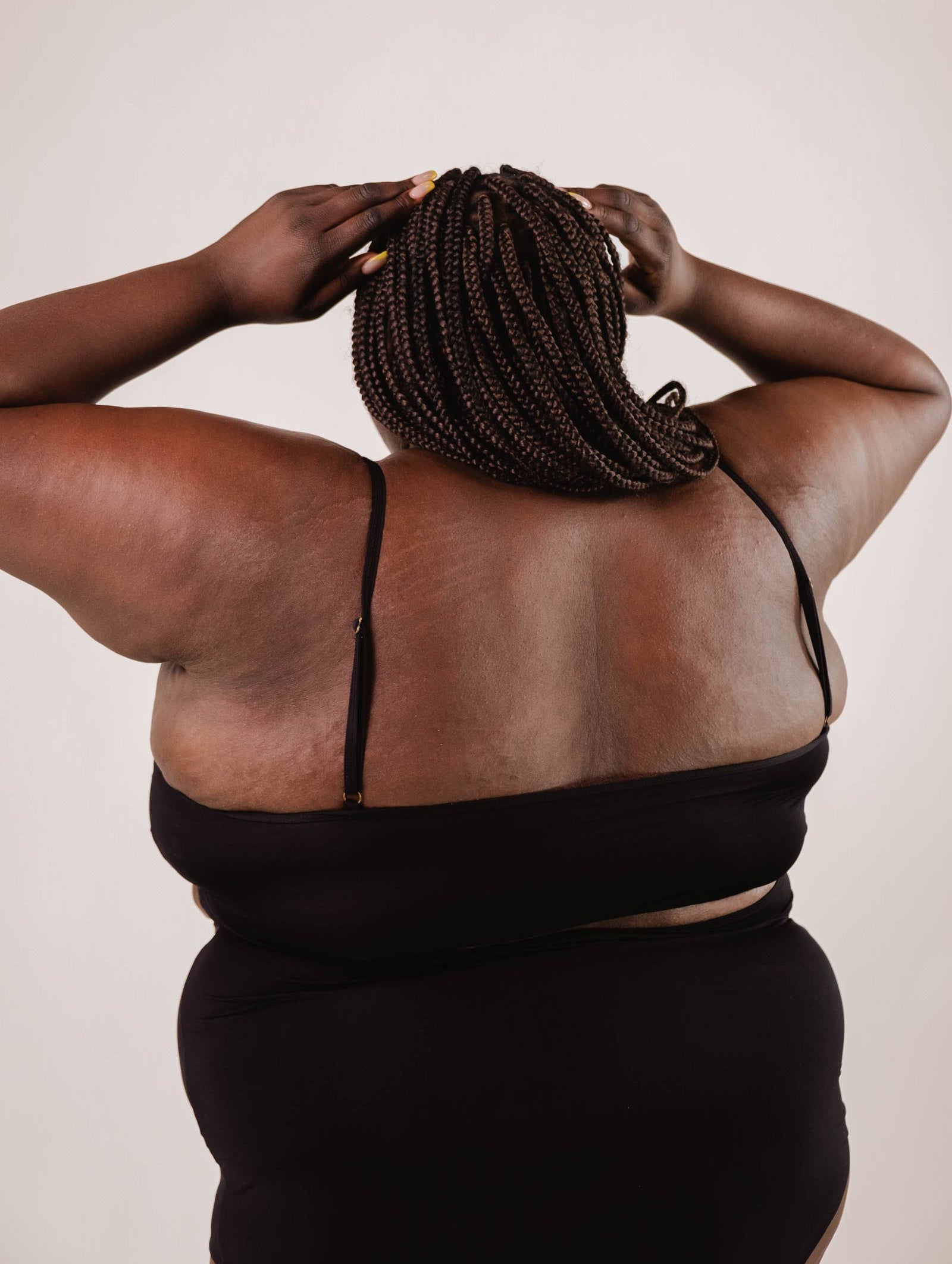 A person wearing a Mimi & August Mango Black Bralette Bikini Top with adjustable straps faces away from the camera, their hands on their head and hair styled in braids.