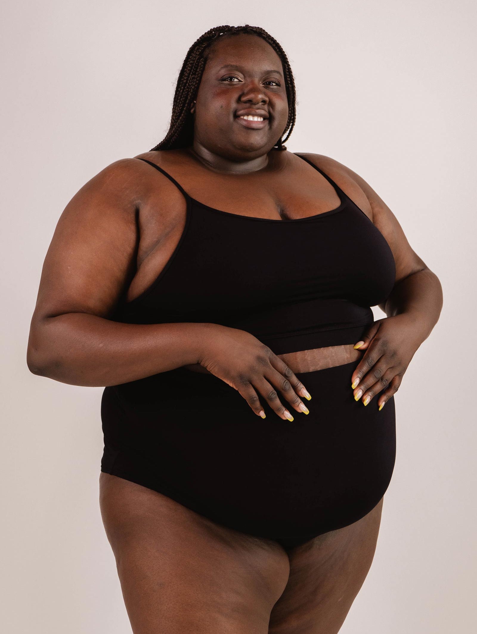 A person wearing a Mimi & August Mango Black Bralette Bikini Top and black shorts stands against a plain background, smiling and looking at the camera. They have braided hair and are holding the edge of their top with their hands, showcasing the adjustable straps that offer excellent support.