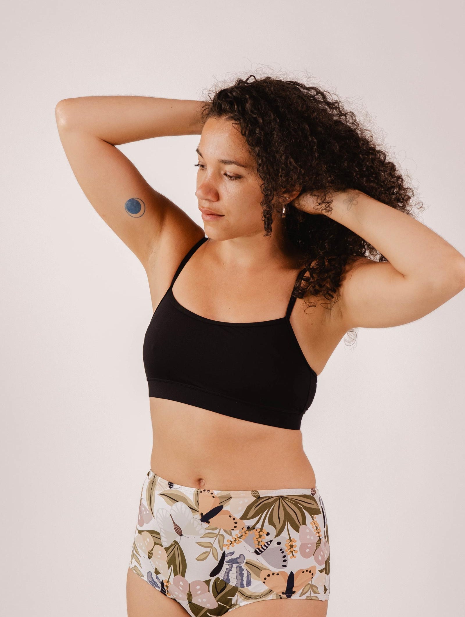 A person with curly hair wearing a Mimi & August Mango Black Bralette Bikini Top and floral-patterned high-waisted underwear, with an armband sensor on their upper arm, stands with arms raised against a plain background.