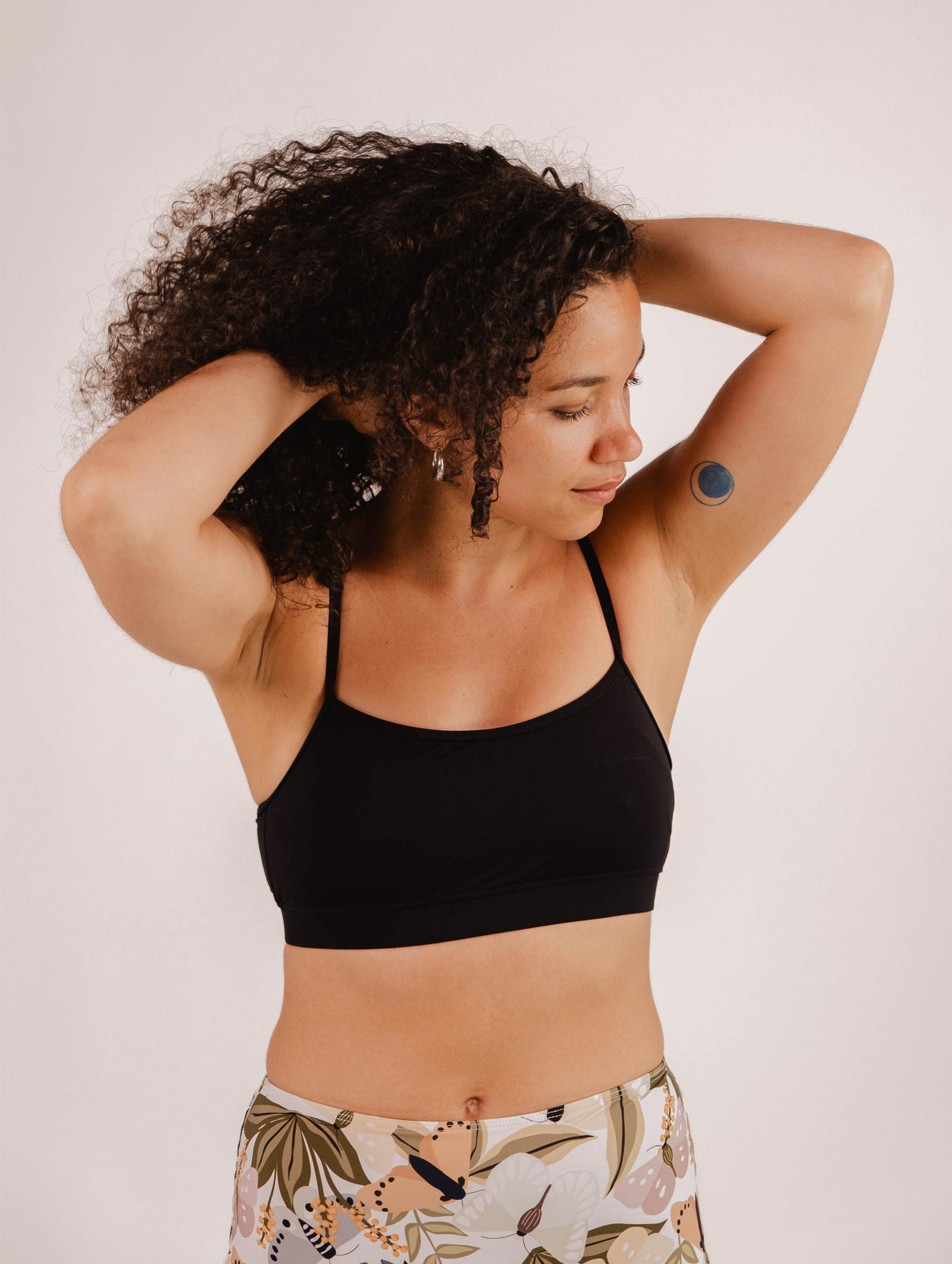 A person with curly hair, wearing a Mimi & August Mango Black Bralette Bikini Top and patterned leggings, adjusts their hair with both hands. A round adhesive patch is visible on their upper arm.
