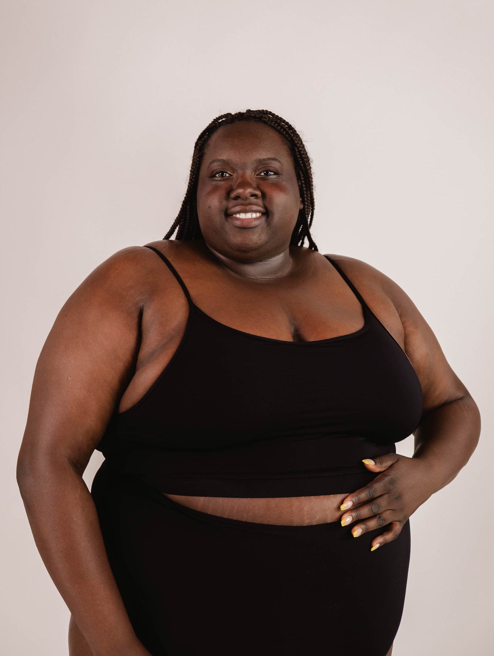 Person stands against a plain background, wearing a Mimi & August Mango Black Bralette Bikini Top with adjustable straps and matching bottoms, smiling with one hand resting on their stomach.