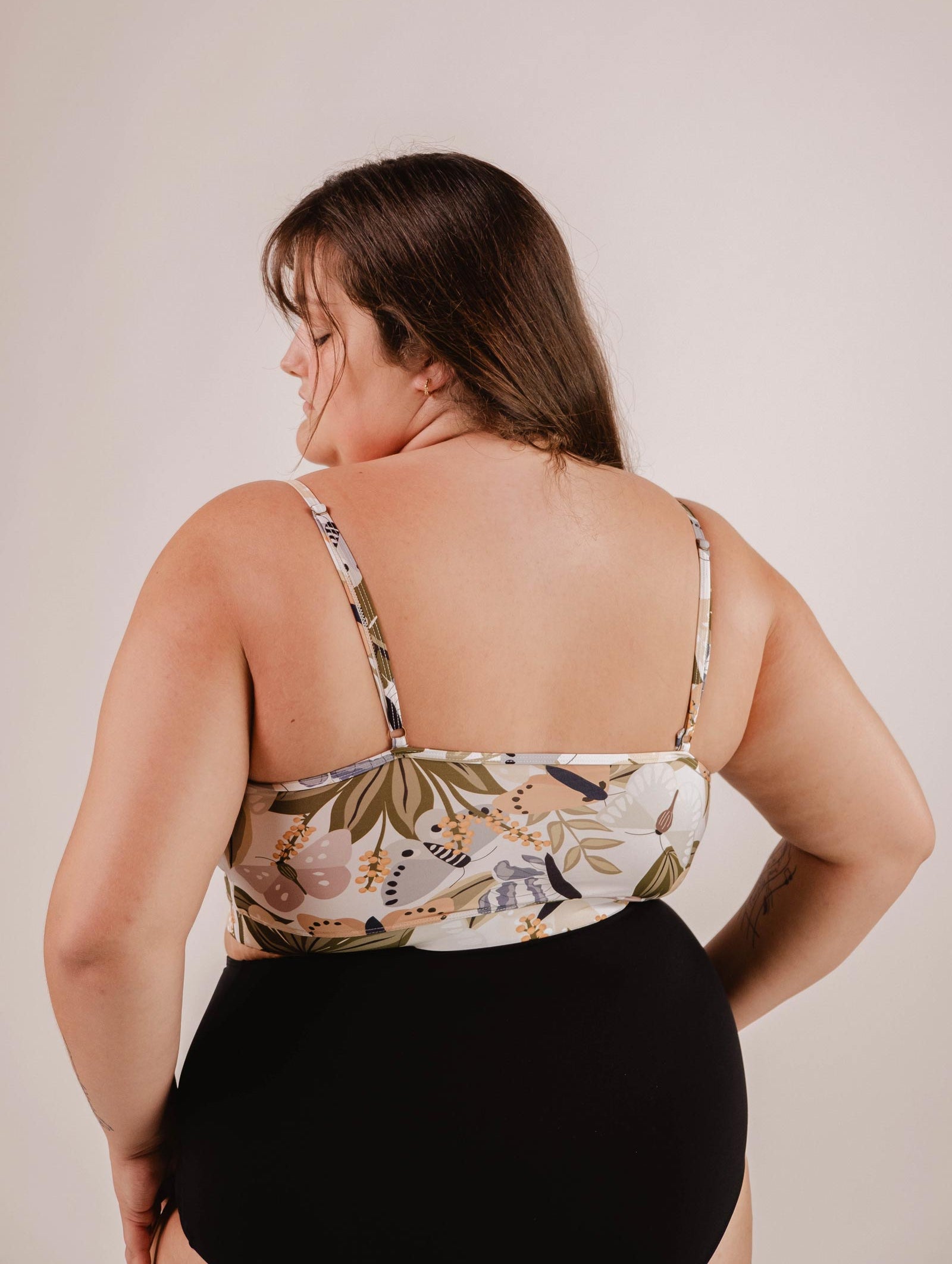 A woman wearing a floral print, sleeveless top and a black skirt or shorts, viewed from the back. The vibrant butterfly patterns on her Mimi & August Mango Mariposa Bralette Bikini Top provide excellent support while adding a touch of elegance to her look.