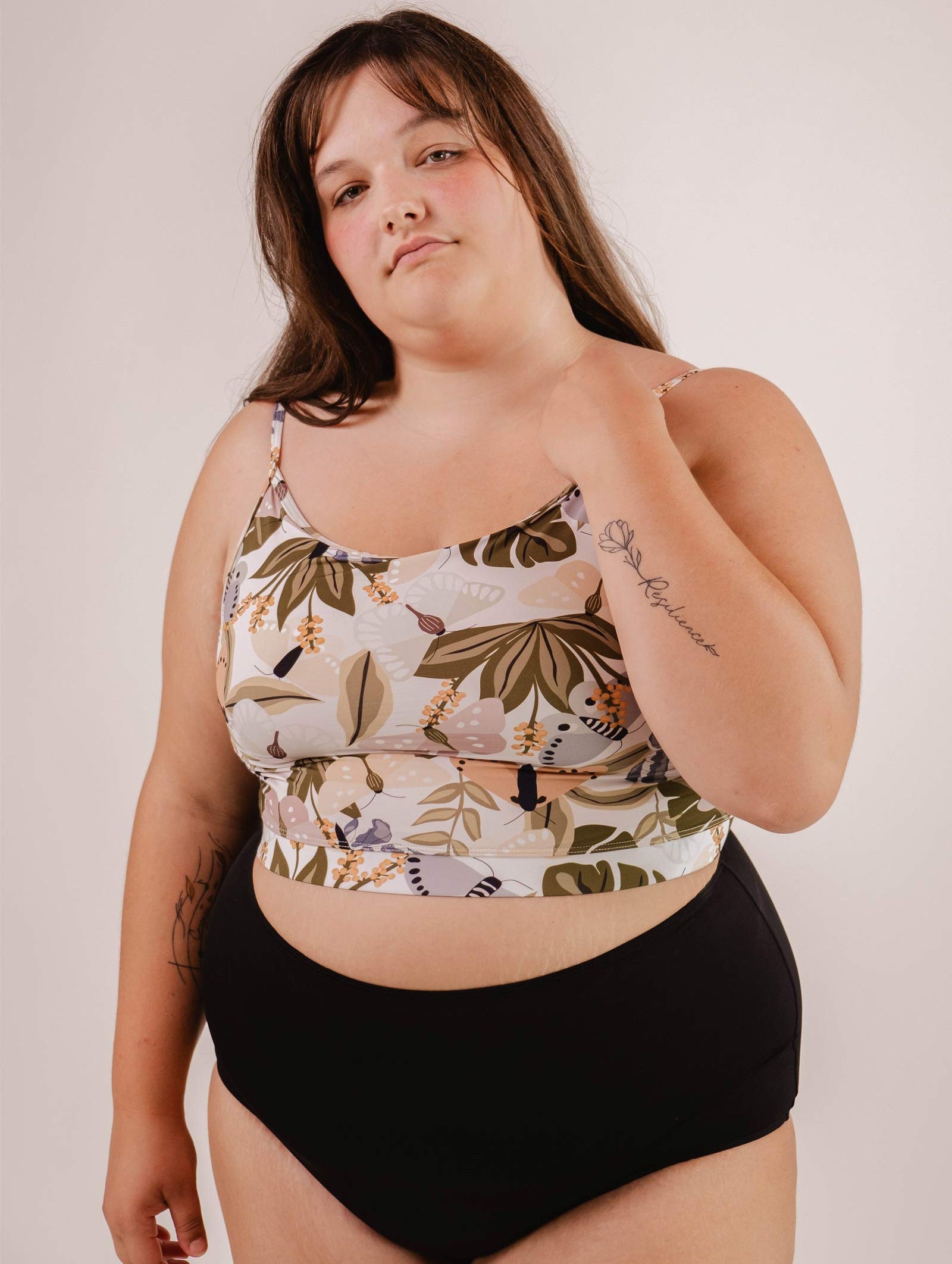 A woman in a Mango Mariposa Bralette Bikini Top by Mimi & August and black bottoms stands against a plain background. She has long hair and tattoos on her arms.