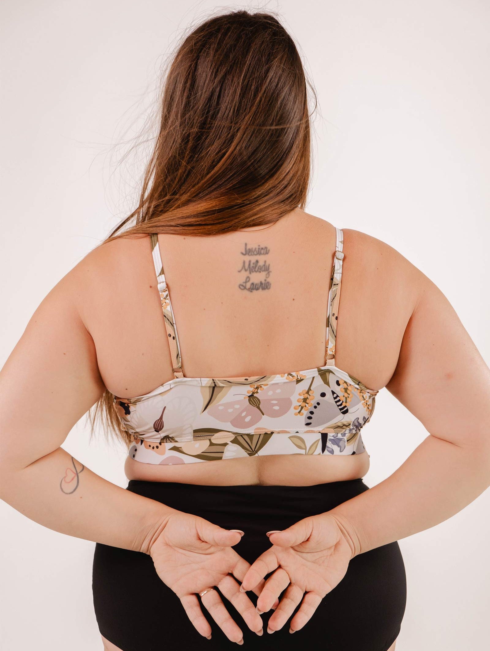 A person with long hair is seen from the back, wearing a Mimi & August Mango Mariposa Bralette Bikini Top adorned with a butterfly and floral print and black bottoms. Their hands are clasped behind their back, revealing a tattoo with names on their upper back.