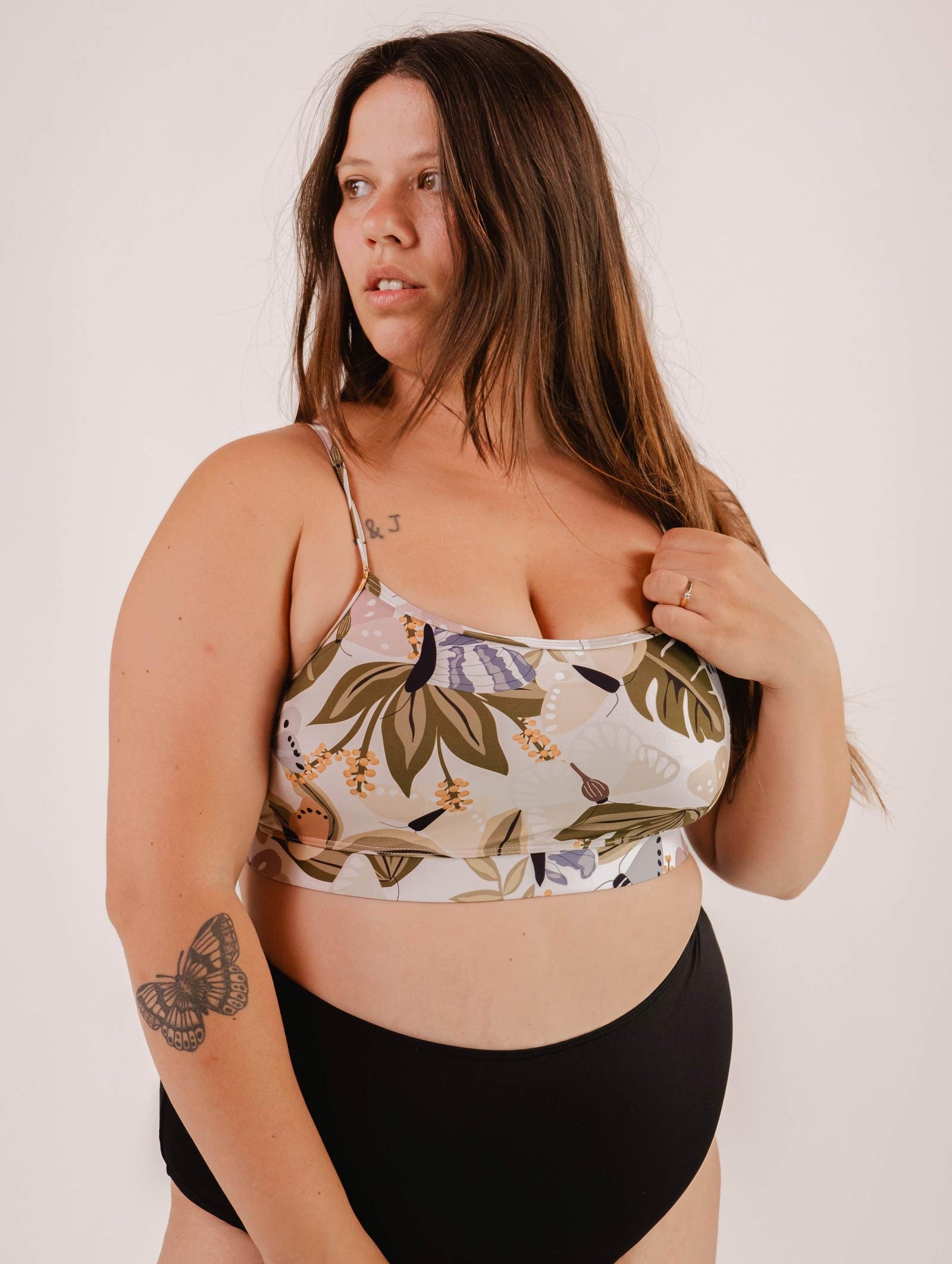Woman with long hair wearing a Mimi & August Mango Mariposa Bralette Bikini Top and black bottoms. She has a tattoo of a butterfly on her upper arm and stands against a plain background, perfectly complementing the butterfly and floral print of her top.