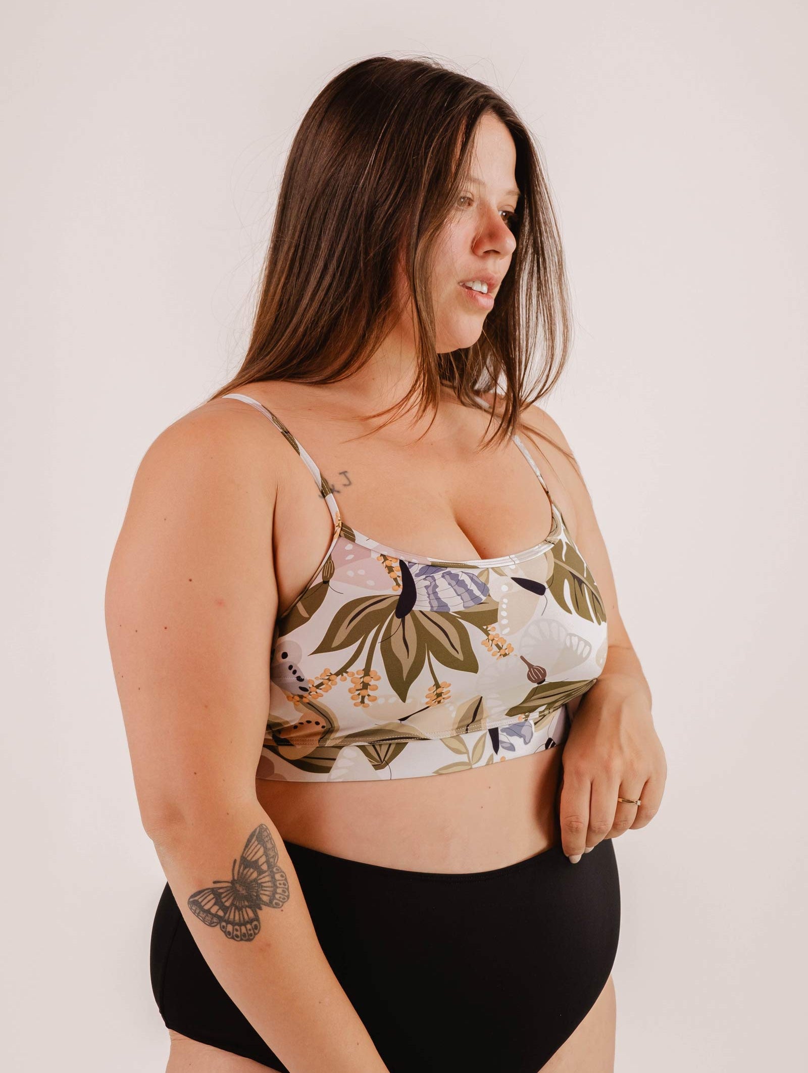 A woman in a Mimi & August Mango Mariposa Bralette Bikini Top and black shorts stands against a plain background. The bikini top, adorned with a butterfly and floral print, provides excellent support. There is a tattoo of a butterfly on her left forearm.