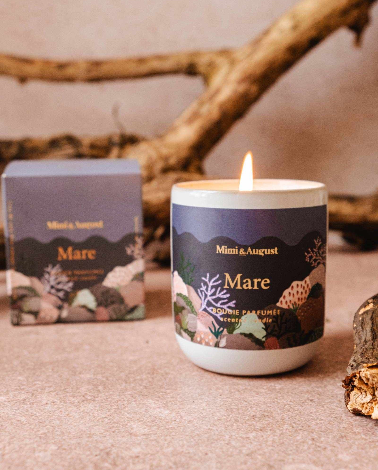 A lit "Mare - Reusable Candle" by Mimi & August, renowned for its calming qualities and ocean scent, placed on a surface with driftwood in the background. A matching product box is shown beside it.