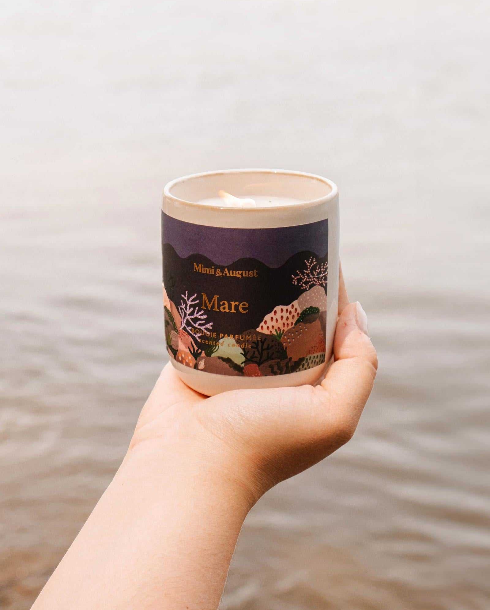 A hand holding a reusable candle with a dark-colored label that reads "Mare - Mimi & August." The background shows a body of water, hinting at its ocean scent and calming qualities.